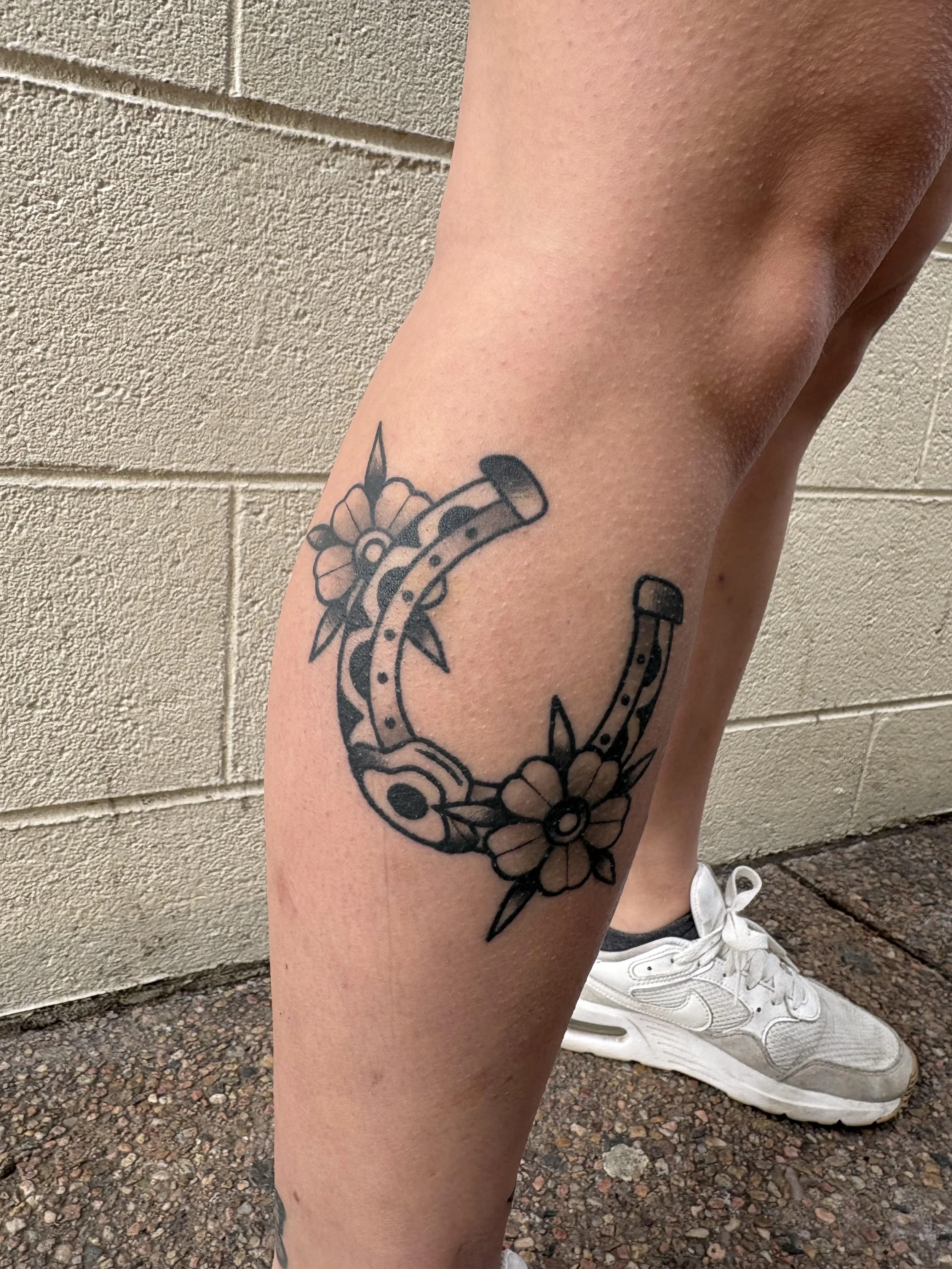 Close-up of a person's leg with a black tattoo of a horseshoe, flowers, and a pin, against a brick wall background, with white sneakers.