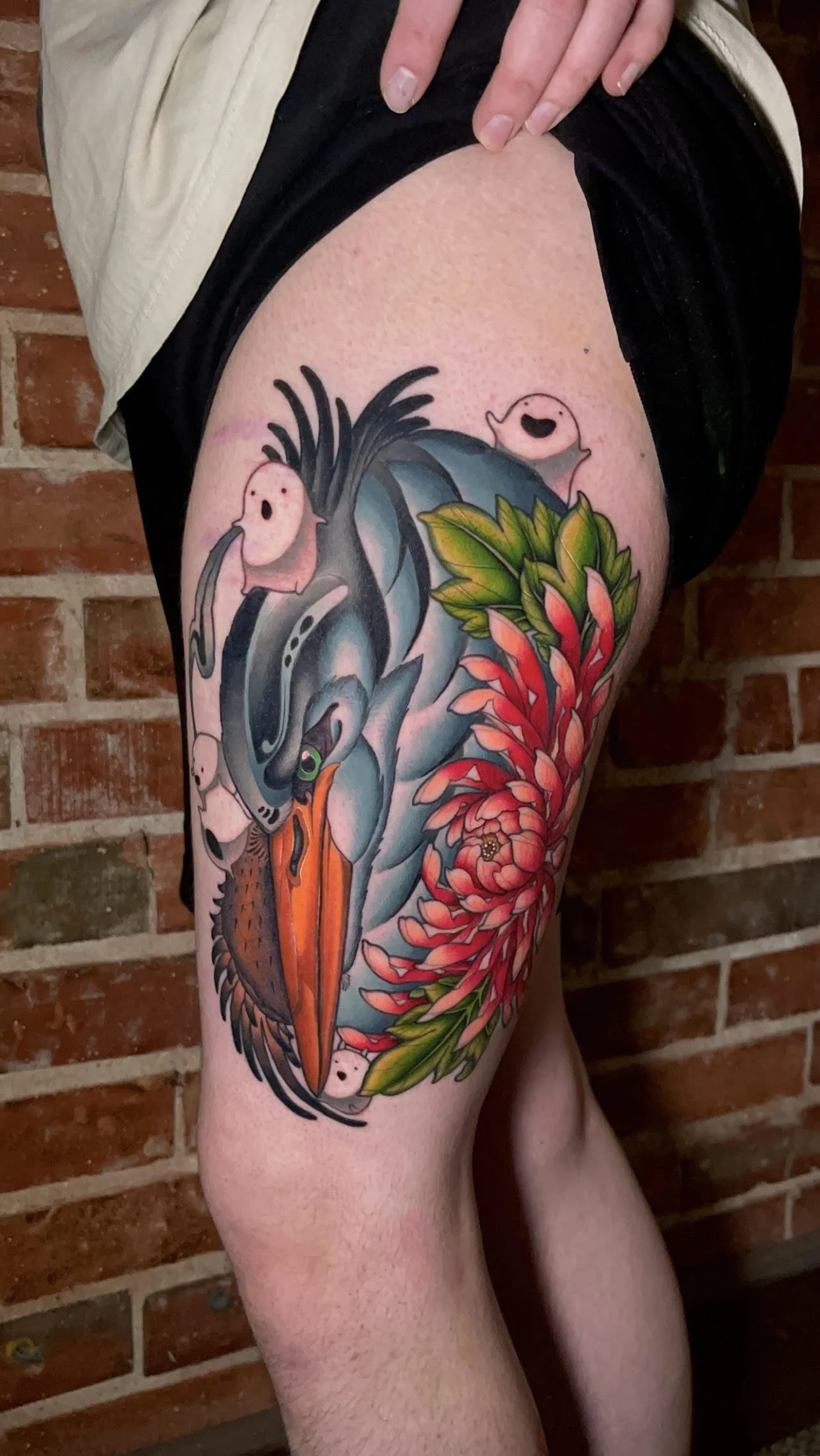 Colorful tattoo of a bird with orange beak and gray wings, surrounded by red flowers and green leaves, on a person's upper thigh.