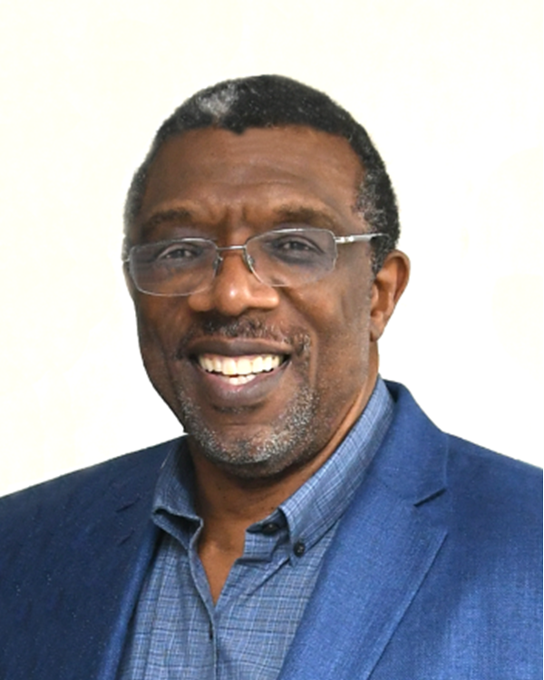 A smiling middle-aged Black man with glasses, wearing a blue suit jacket and blue shirt, standing against a light background.