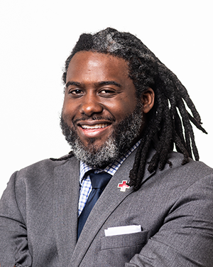 A man with dreadlocks, wearing a gray suit and tie, smiling, with a health-related lapel pin, against a white background.