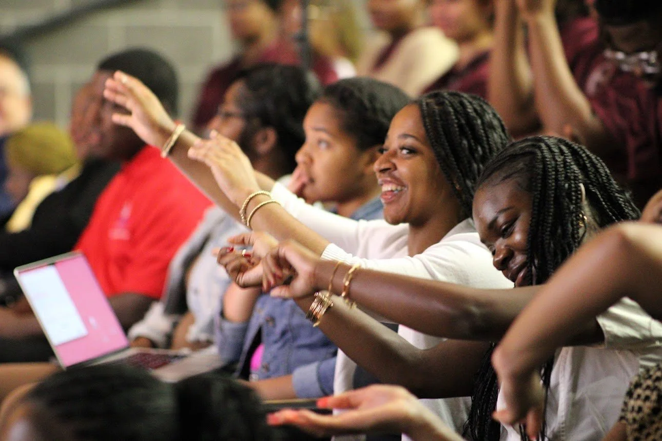 A group of women sitting in an audience, smiling and laughing, with some raising their hands and others looking at their laptops, in a lively setting.