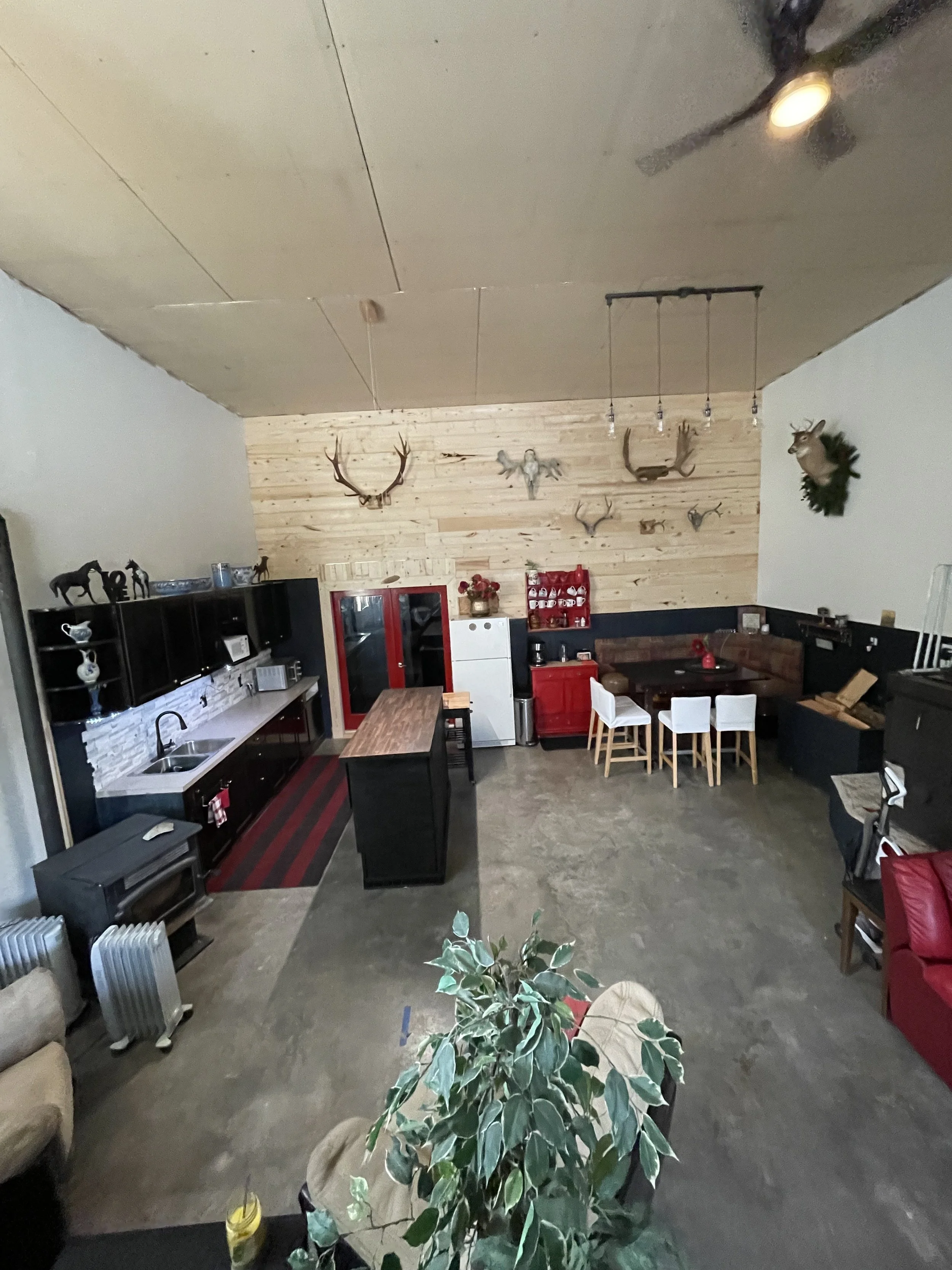 Sixteen‑foot ceilings rise above you like the inside of a timbered retreat, the ceiling fan circling lazily in the warm glow of Edison bulbs. Their soft light spills across the room, catching the edges of the wood stove — the heart of the space — whe