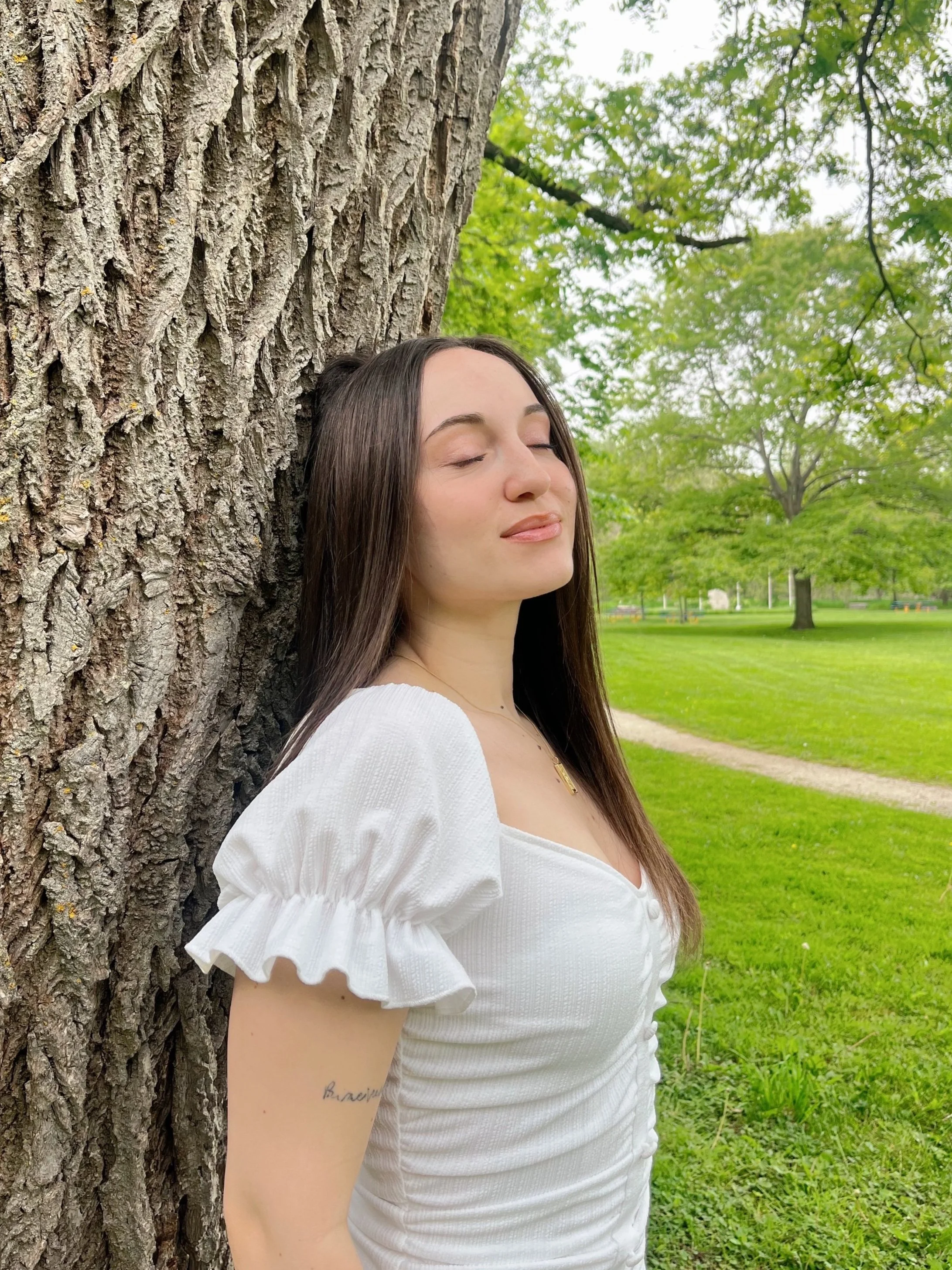 A young woman with long dark hair, wearing a white dress with puffed sleeves, leaning against a large tree with eyes closed in a park with green grass and trees.