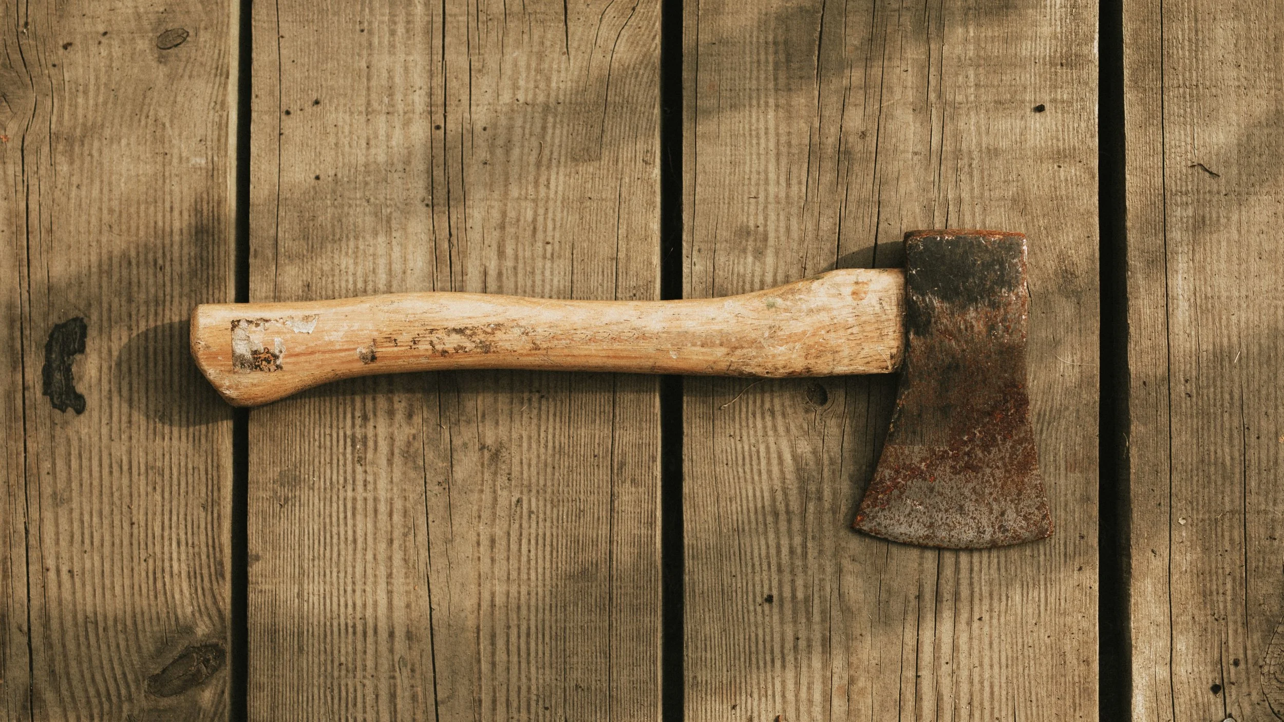 An old axe with a wooden handle and rusted metal head lying on a wooden surface.