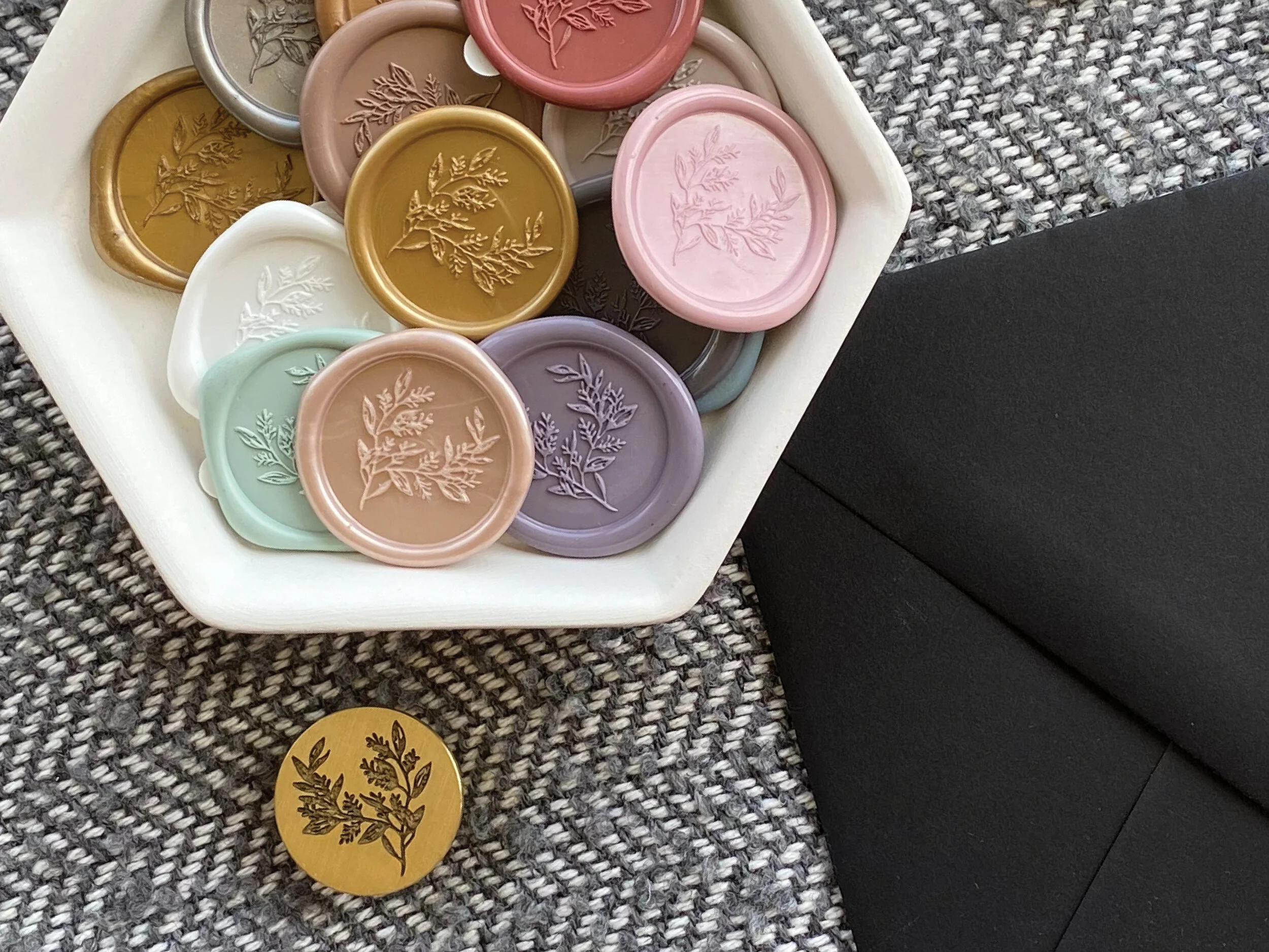 A white hexagonal plate holding an assortment of colorful wax seals in pink, purple, white, gold, and gray with embossed botanical designs, with one gold seal on a gray, textured surface nearby.