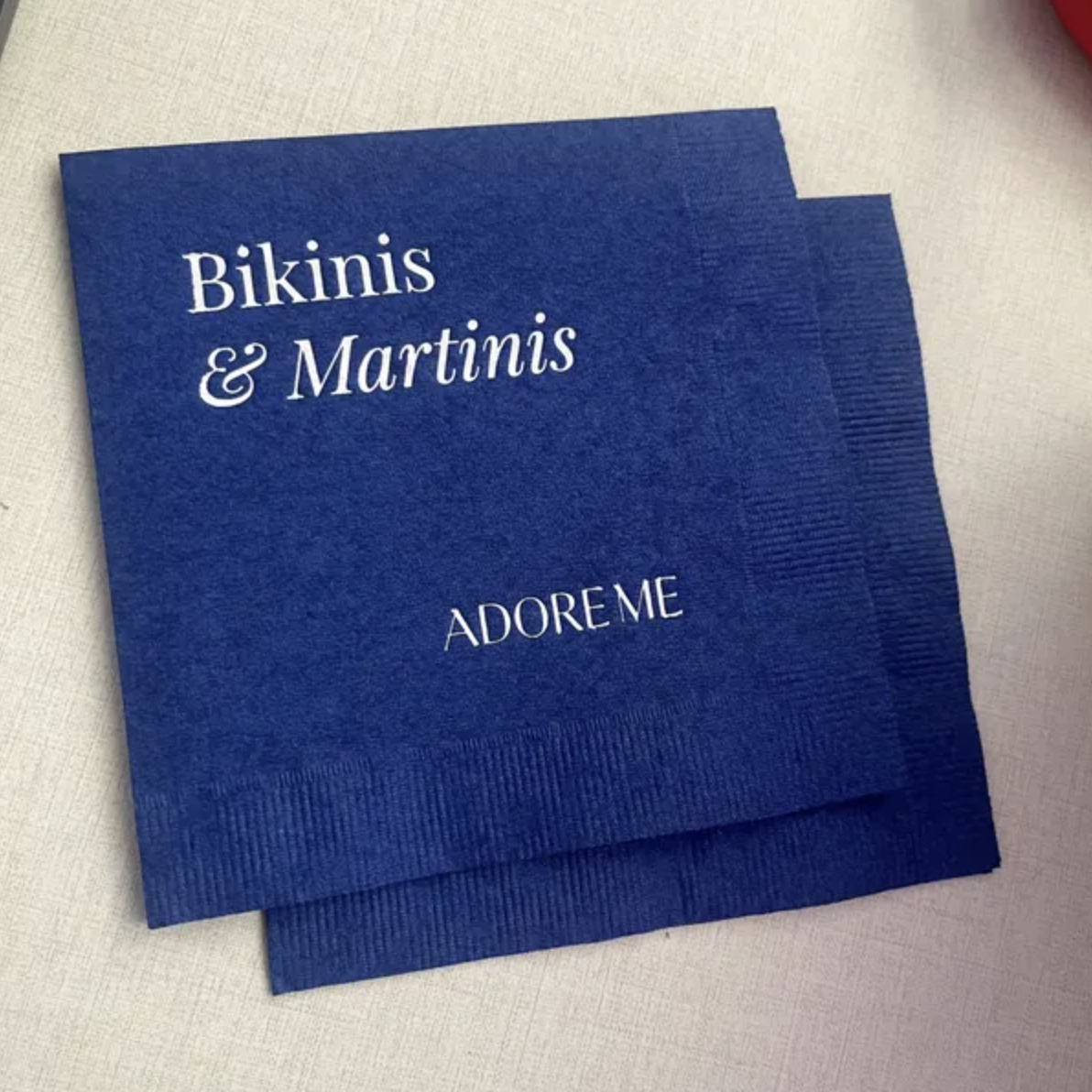 Two blue napkins with white text that read 'Bikinis & Martinis' and 'ADORE ME' on a white table.