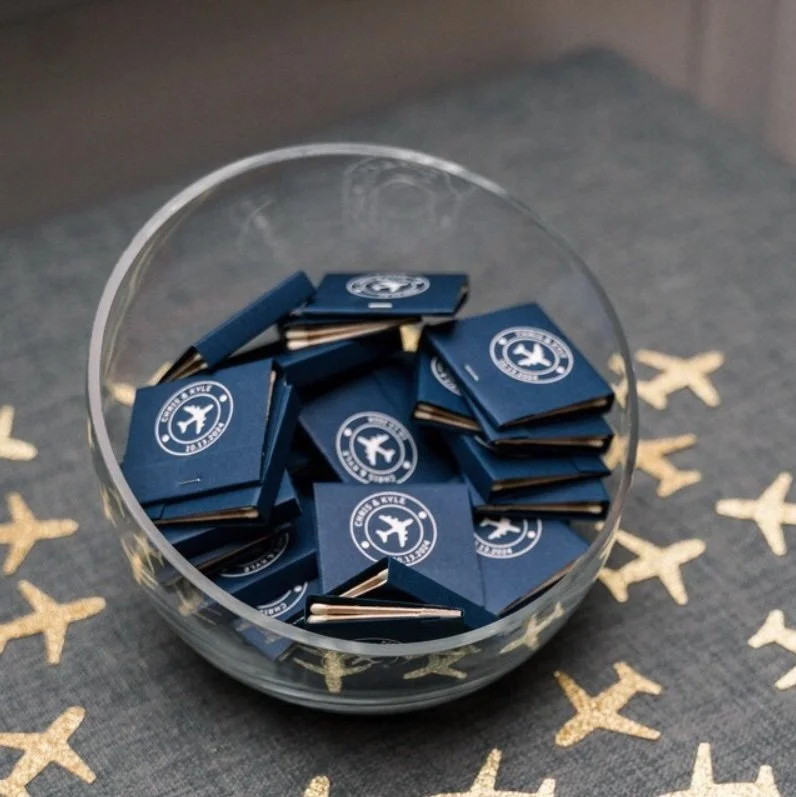 A glass bowl filled with small, blue, rectangular objects bearing a white logo of an airplane surrounded by the words 'Enjoy the Style.' The bowl is placed on a gray fabric surface decorated with cream-colored airplane patterns.