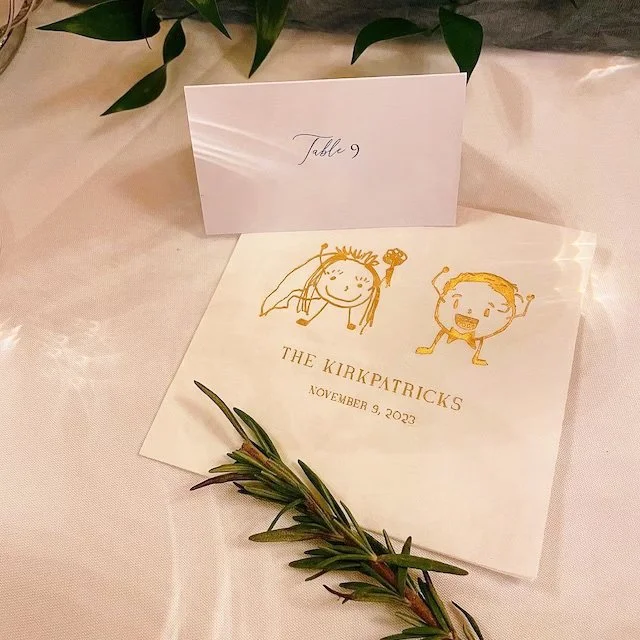 A wedding program with cartoon illustrations of a bride and groom, labeled 'The Kirkpatricks' and dated November 9, 2023, on a table with a white tablecloth. There is a small place card labeled 'Table 9' and a sprig of greenery.