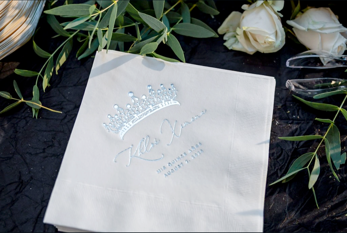A white napkin with a silver embroidered crown and text that reads "Kale Xtreme" and "MIS QUINCE AÑOS, AUGUST 2, 2023" on a black surface surrounded by green leaves, white flowers, and clear plastic utensils.