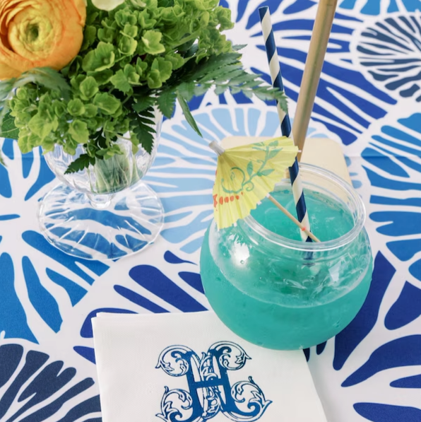 Blue cocktail with ice in a glass jar, garnished with a small paper umbrella and two striped paper straws, placed on a blue and white patterned tablecloth with a floral centerpiece including green flowers and leaves in a glass vase.