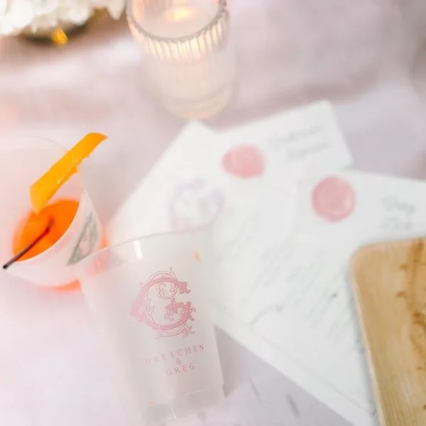 Frost-Flex Shatterproof plastic cup with pink and green monograms logo, an orange cocktail pick with fruit garnish, and menus with pink wax seals