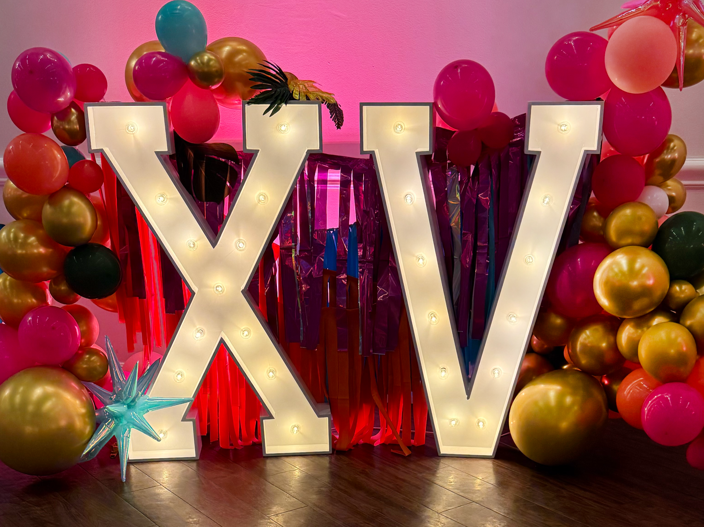 Large illuminated marquee letters spelling 'X' and 'V' surrounded by colorful balloons in pink, gold, and black, with pink and purple streamers and a small turquoise star-shaped balloon decoration.