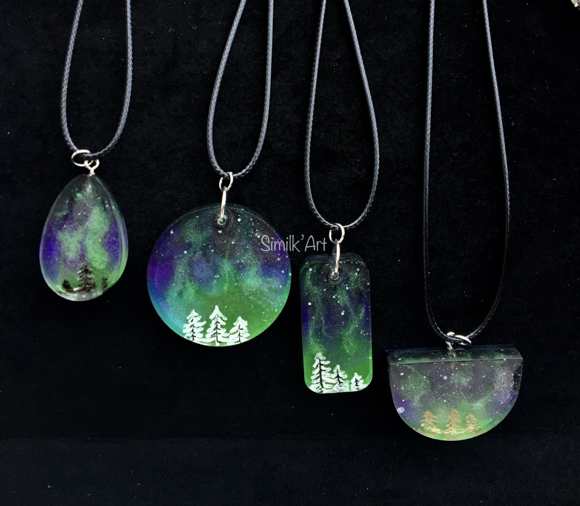 Four handcrafted pendants with painted night sky and northern lights scenes, featuring trees at the bottom, hanging on black cords.