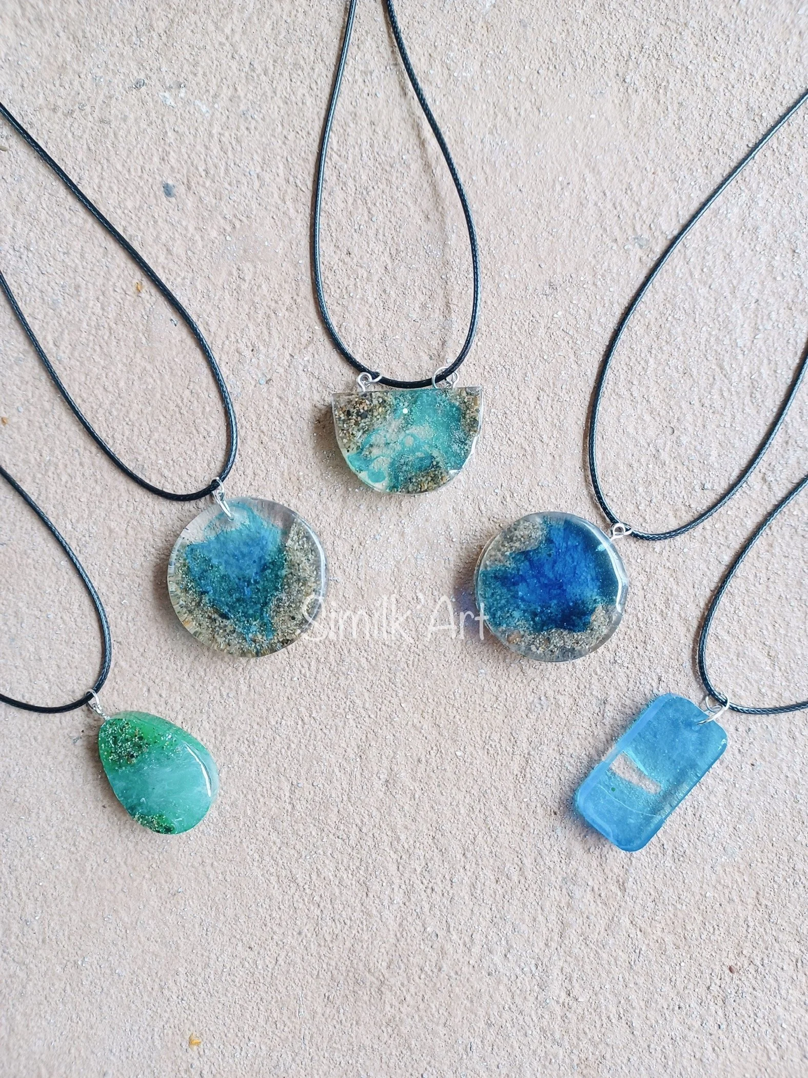 Five ocean themed necklaces with blue and green resin pendants on a beige background.