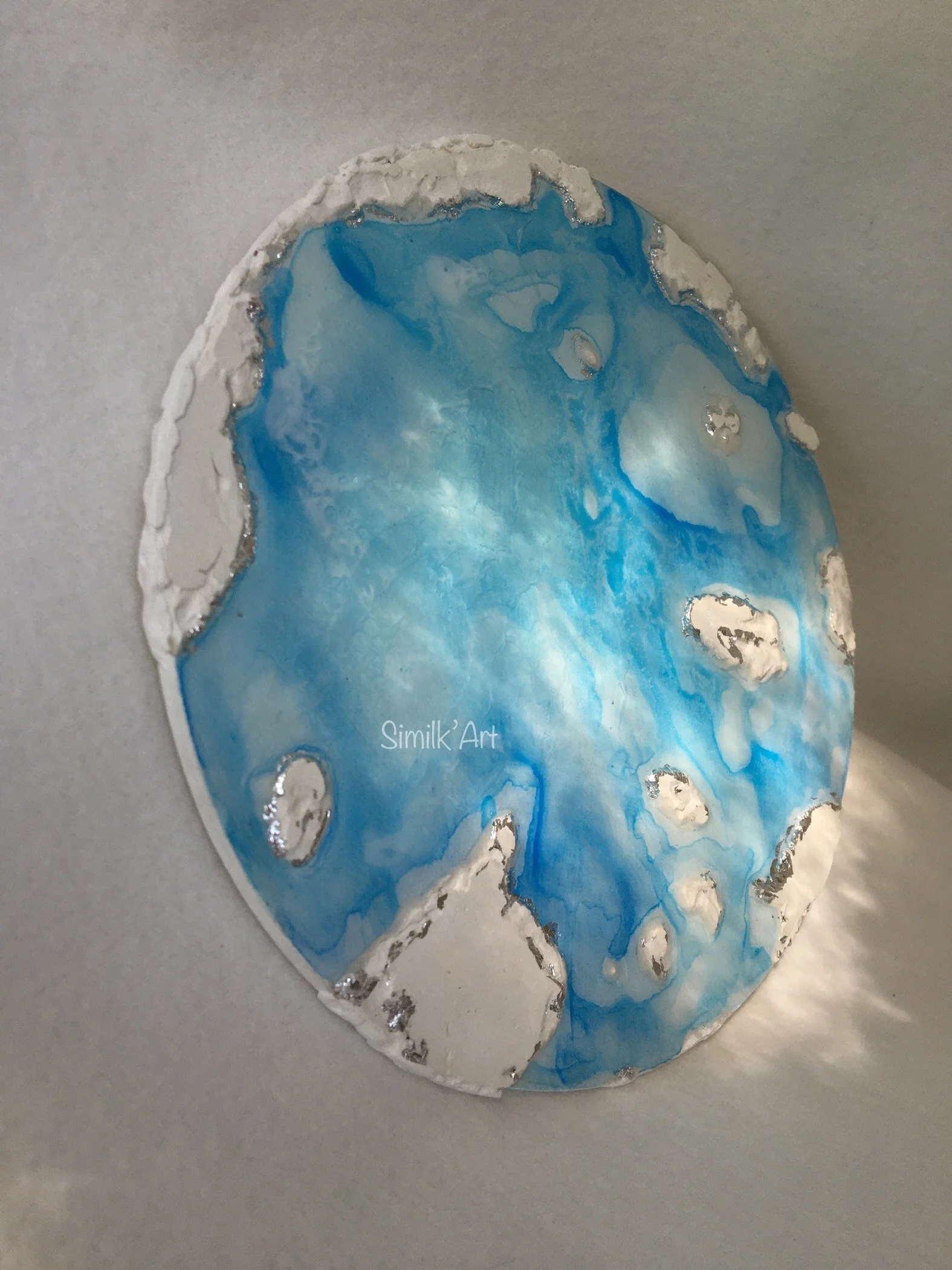 A circular abstract Mixed Media art piece made with plaster, resin, blue alcohol ink pattern and waves. It contains silver accents around some sides of the plaster.