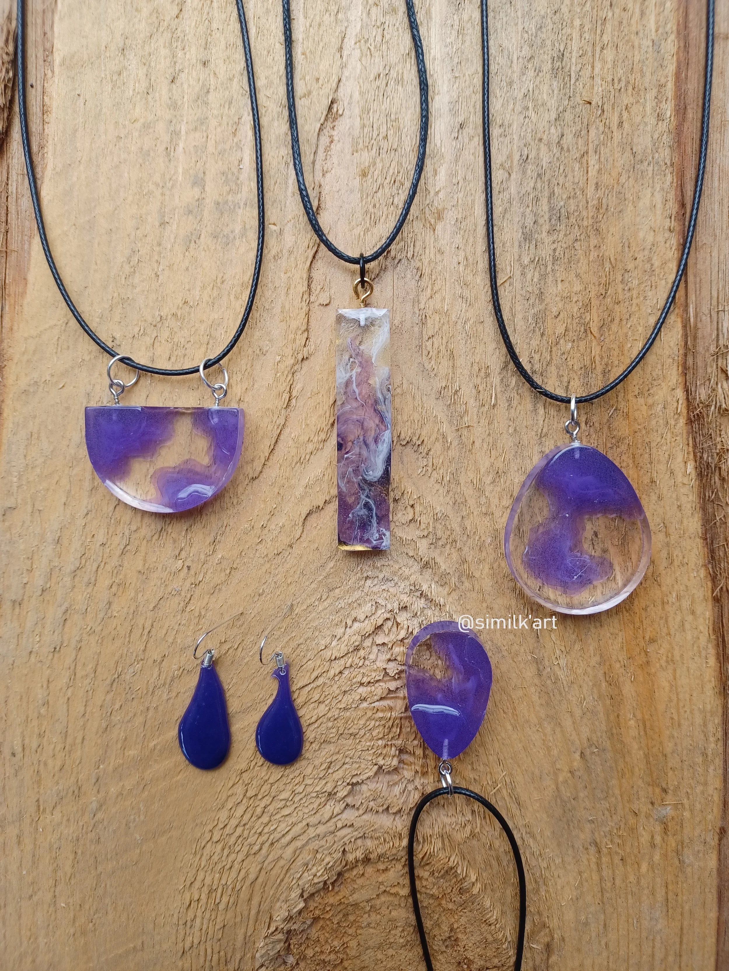 Collection of purple jewelry necklaces and earrings displayed on a wooden background, featuring various pendant shapes including semi-circle, rectangle, teardrop, and oval.