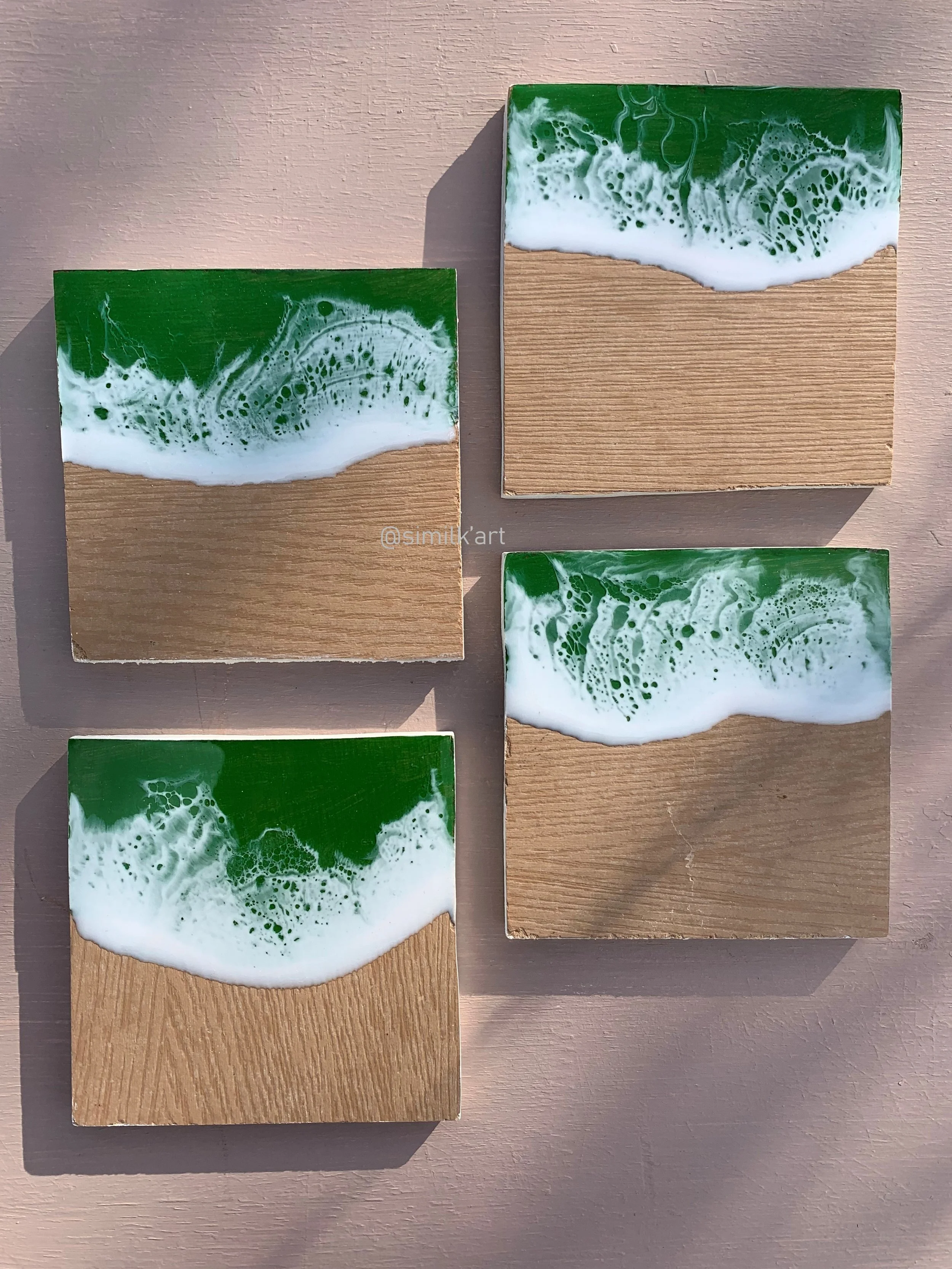 Four square wood plaques with green and white resin art resembling ocean waves, arranged on a pink surface.
