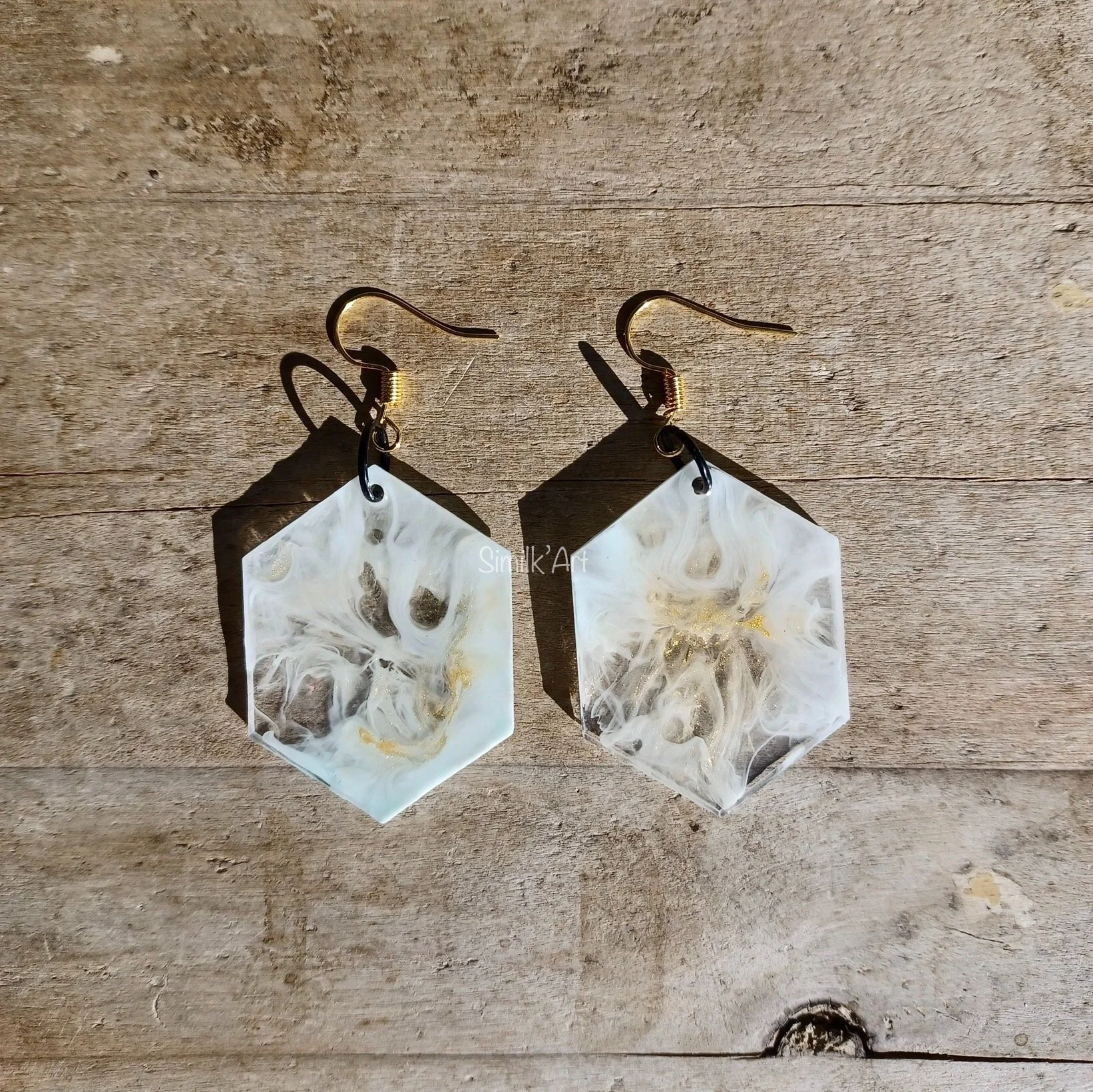 Pair of geometric resin earrings with white, clear, and gold swirling patterns, hanging on metal hooks against a wooden background.