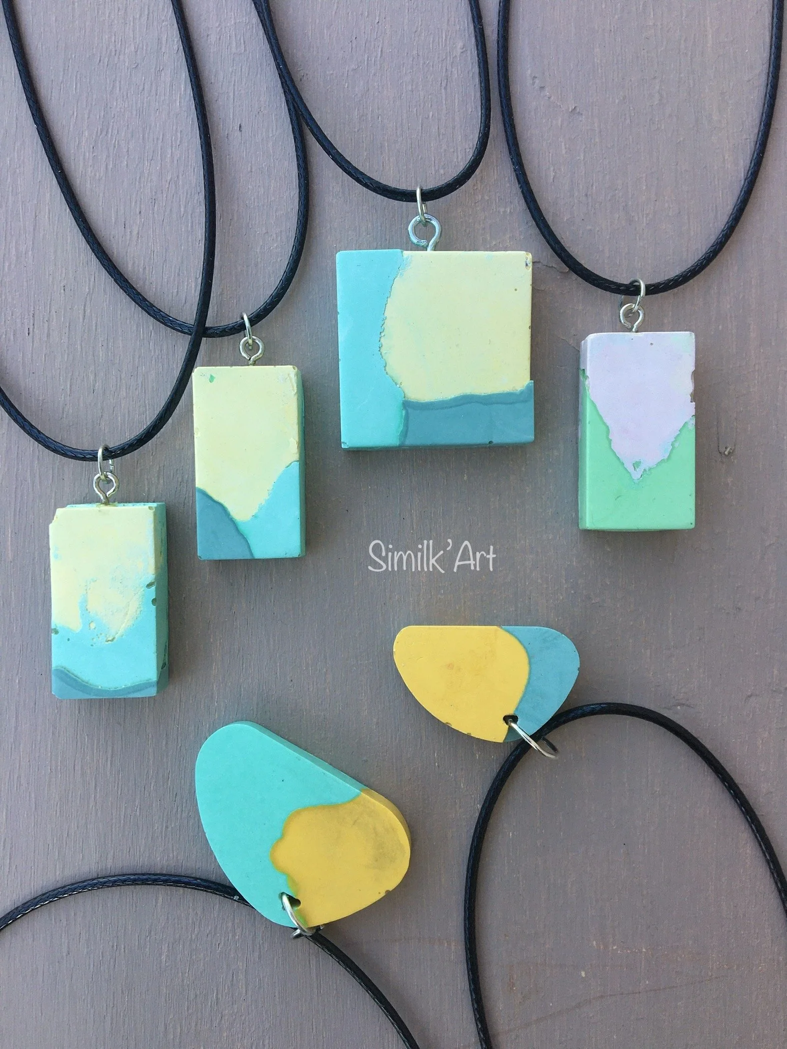 Colourful handmade jewelry made of Jesmonite including rectangular, round and square necklaces with abstract blue, yellow, and pink designs on black cords, displayed on a gray background.