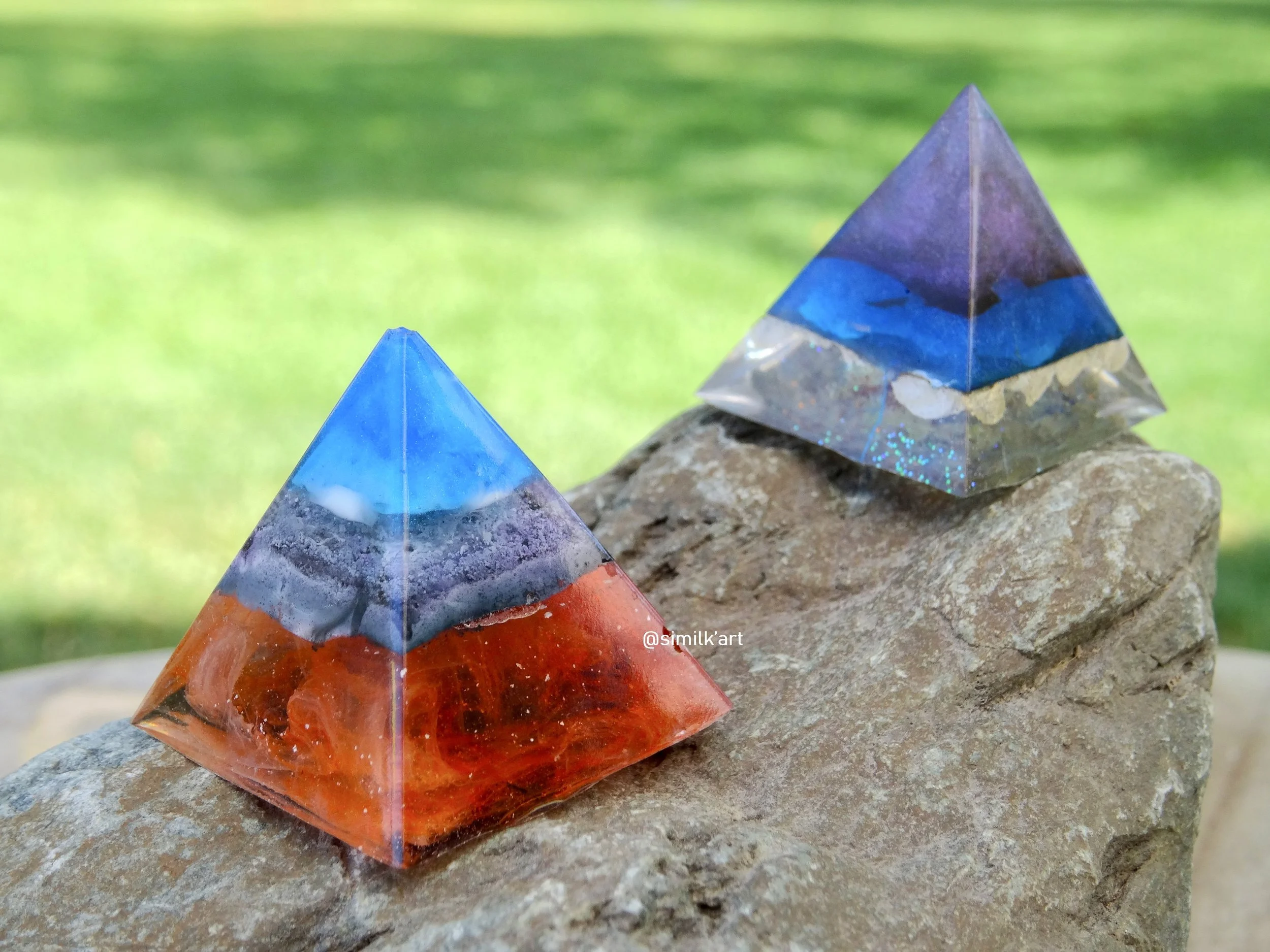 Two colourful artistic pyramid sculptures, one with red, blue, and purple hues, and the other with purple, blue, and hints of glitter, are placed on a rough stone surface outdoors with a green grassy background.