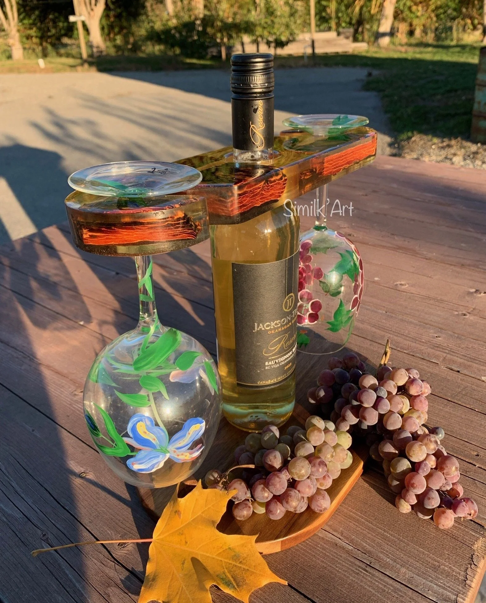 A wine caddy made of Resin & wood supporting a bottle of white wine on a wooden table, with two hand-painted wine glasses, bunches of grapes, a yellow maple leaf. The scene is outdoors with trees and a sunset in the background.