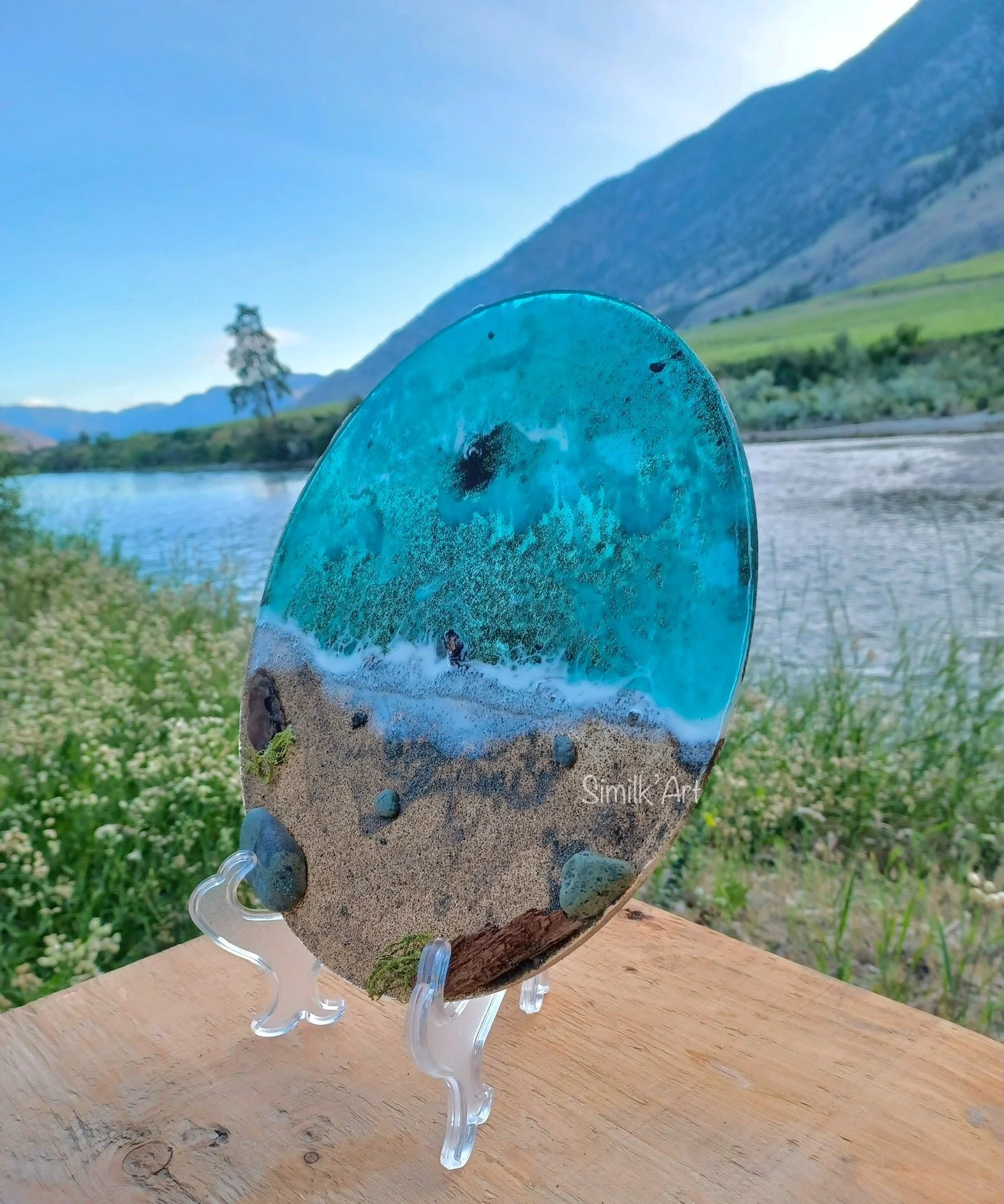 A decorative emerald green resin art piece depicting a beach scene with ocean waves, sand, rocks, and a clear blue sky, displayed on a stand outdoors with mountains and a river in the background.