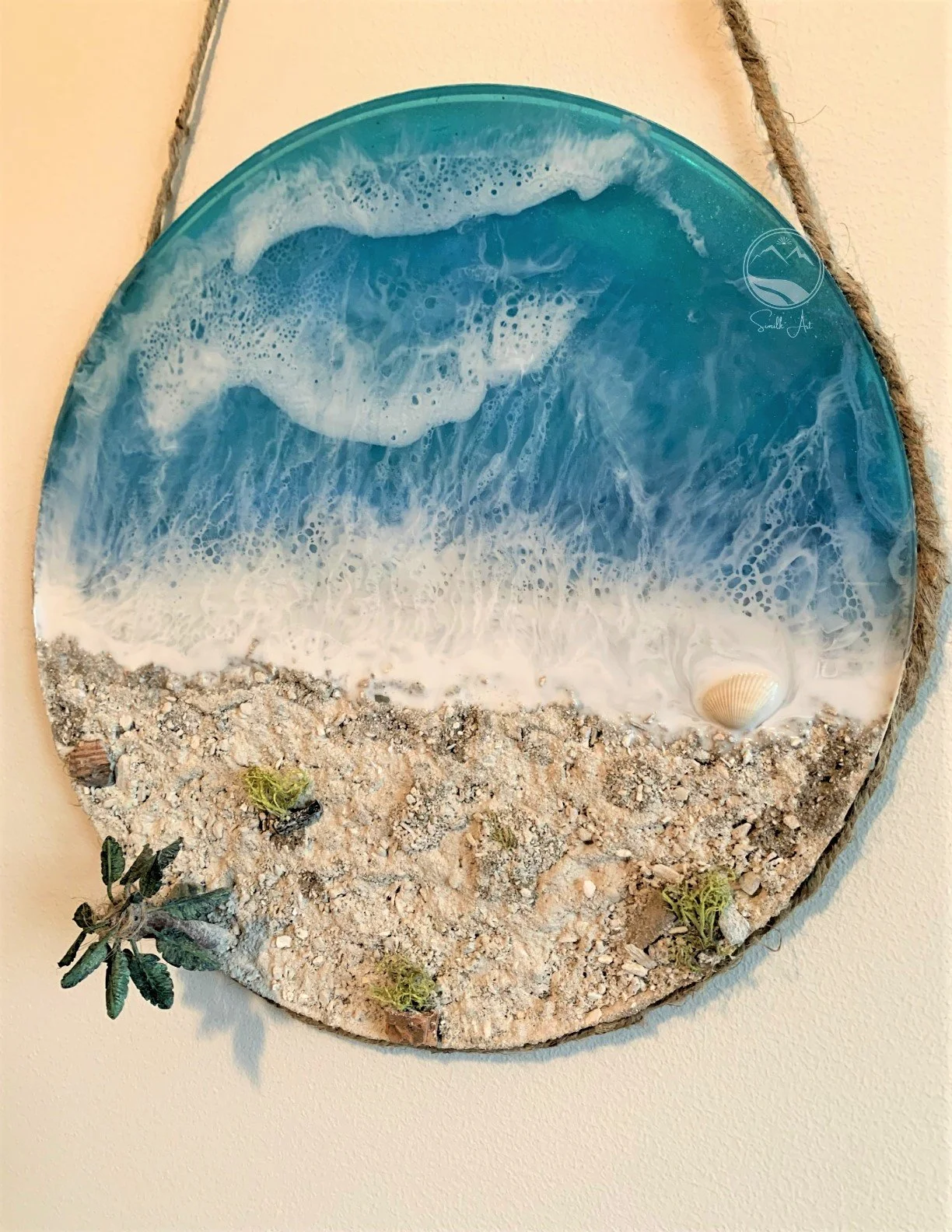 Decorative art piece depicting an ocean scene with waves, sand, shells, and small plants, mounted on a wall and framed with a wooden circle.