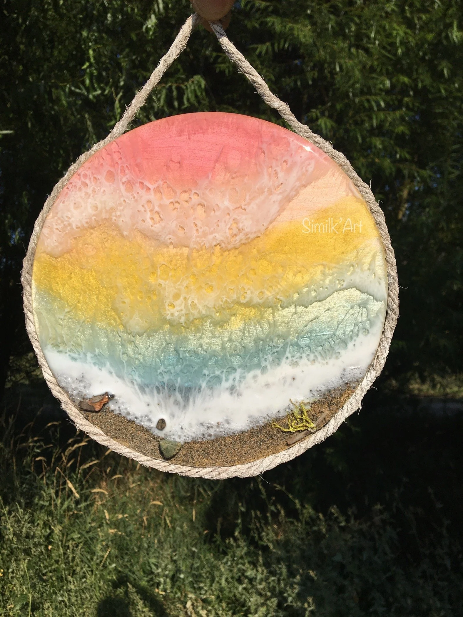 A round piece of art resembling a sunset over the ocean, with red, yellow, green, and blue colors, is hanging outdoors against a background of green trees. The art is on a textured surface with a rope around its edge for hanging.