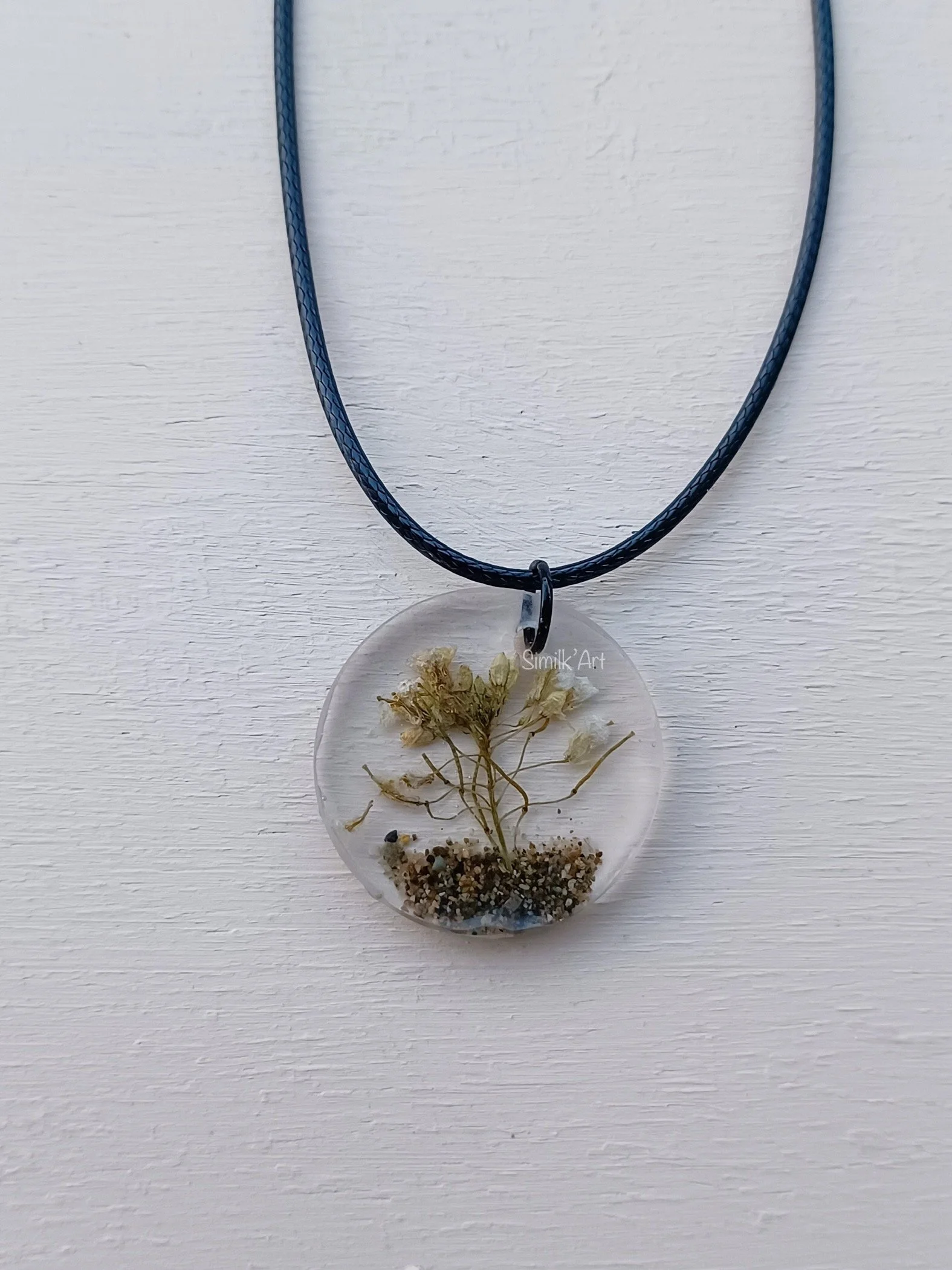Pendant necklace with a clear resin circular charm containing dried flowers and soil, hanging from a black cord.