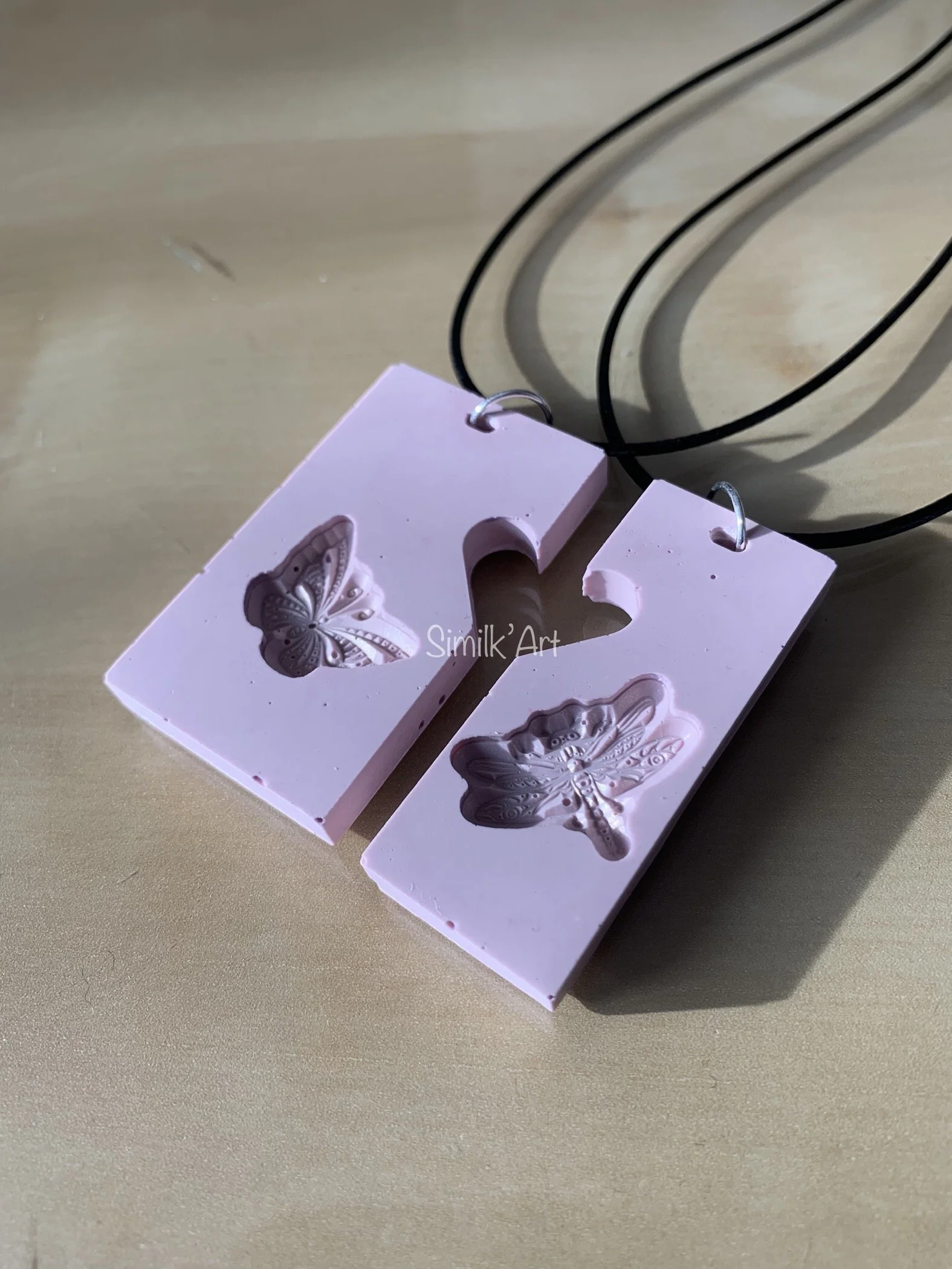 Pink Jesmonite earrings with butterfly and dragonfly designs, hanging from black cords, placed on a light wooden surface.