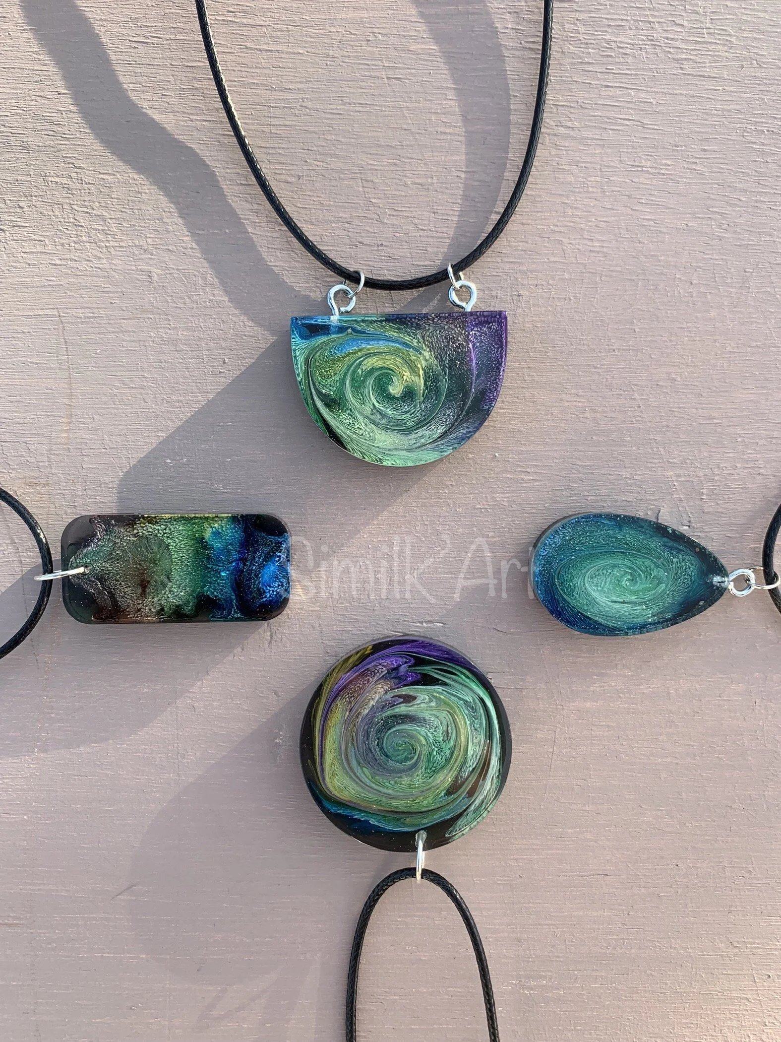 Collection of colorful, swirling resin pendants simulating the Petri effect with black cords on a pink wooden surface.