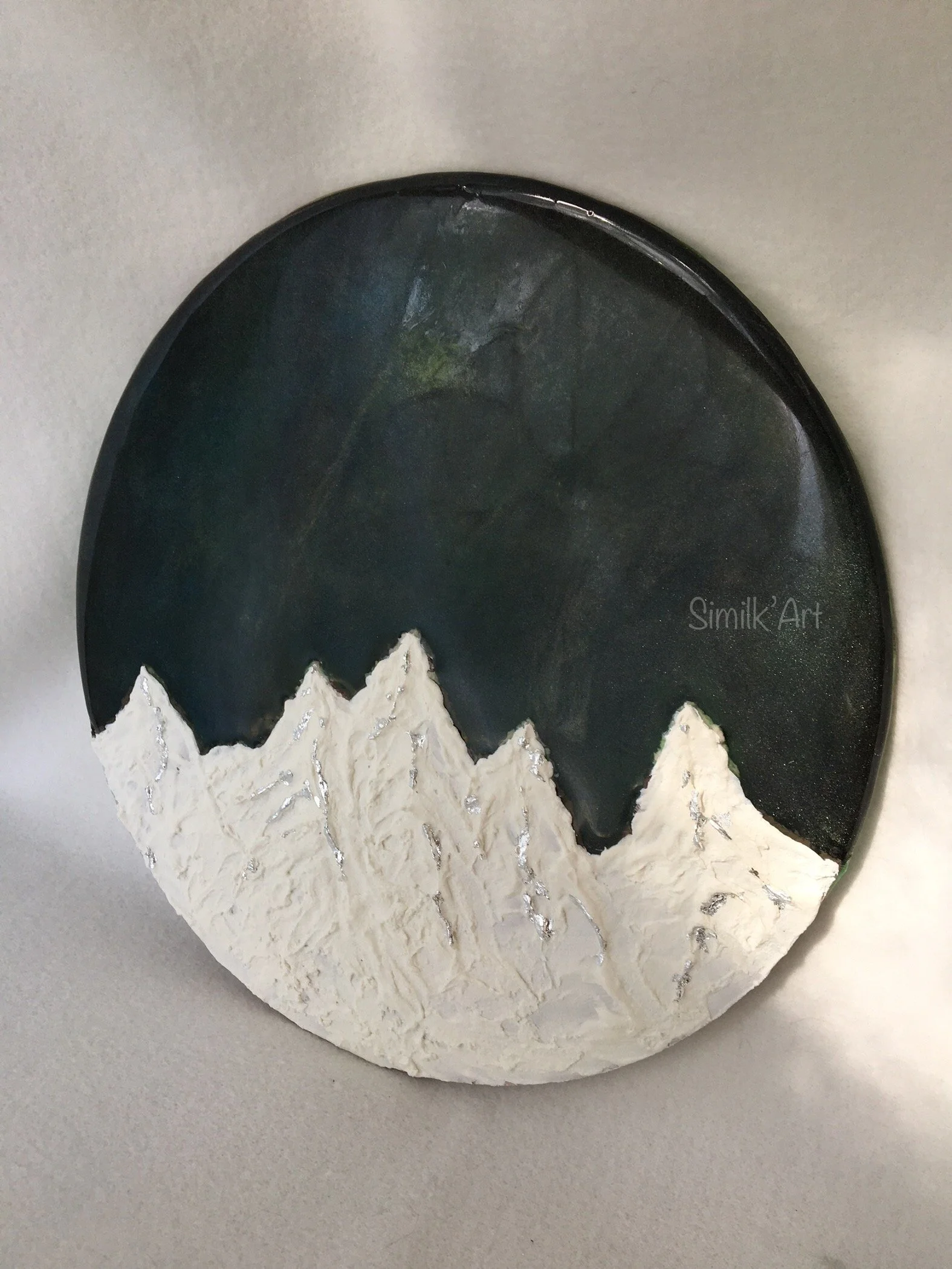 A circular glow in the dark art piece depicting snow-covered mountains against a dark sky. The mountains are textured and painted white with silver accents, and the sky is painted in dark shades with some subtle hints of green and blue.