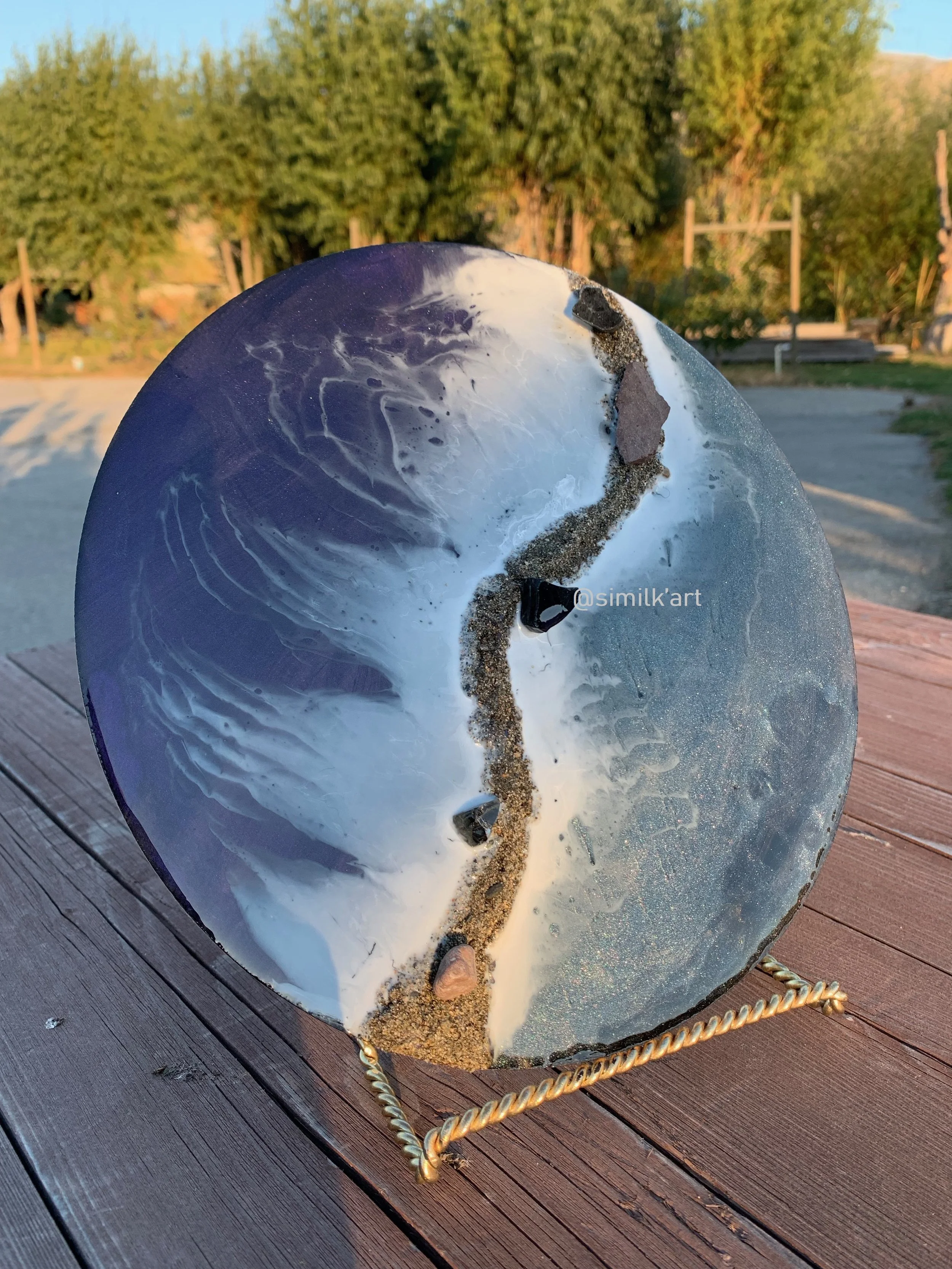 A round ocean themed resin art piece with black, white, and purple patterns, embedded with small fire pit crystals and sand, displayed on a wooden surface with a landscape of trees in the background.