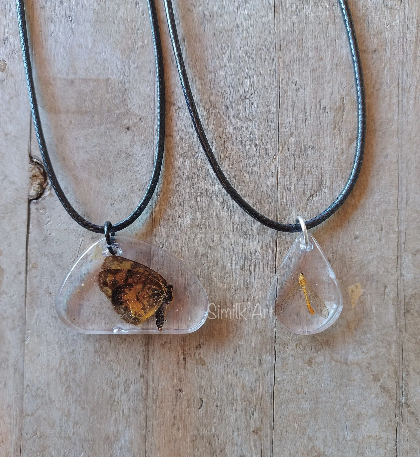 Two necklaces with resin pendants, one contains a butterfly and the other contains a dragonfly, hanging against a wooden background.