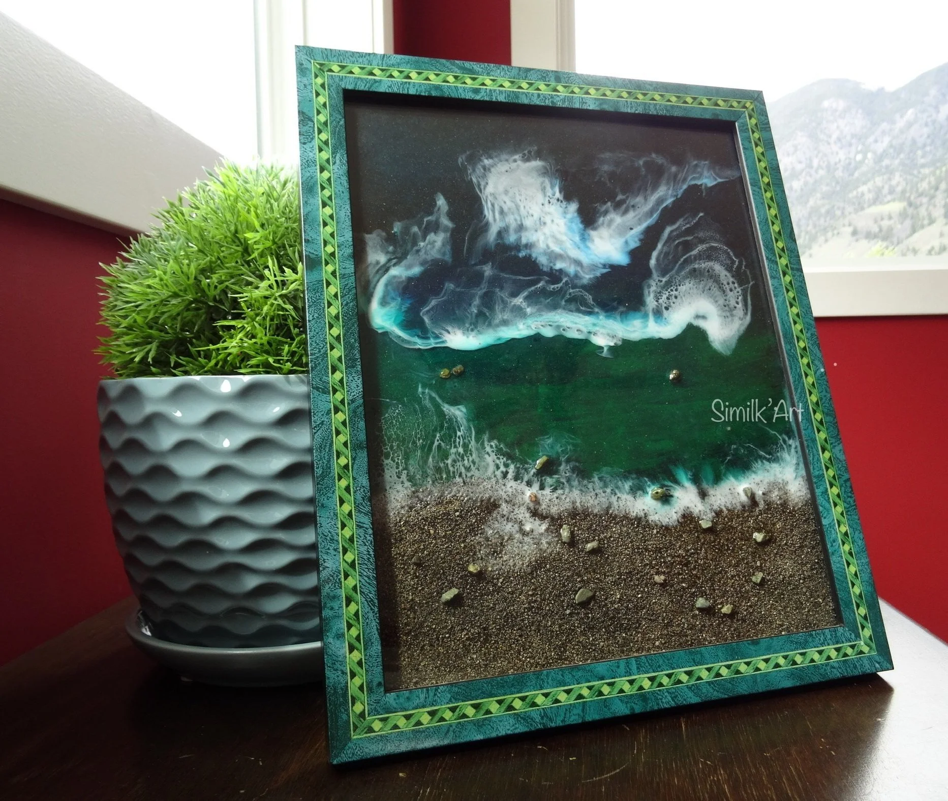 Framed 3D ocean artwork with waves, beach, and sea foam, placed on a wooden surface next to a potted green plant with textured gray pot, in front of a red wall and window with mountains outside.