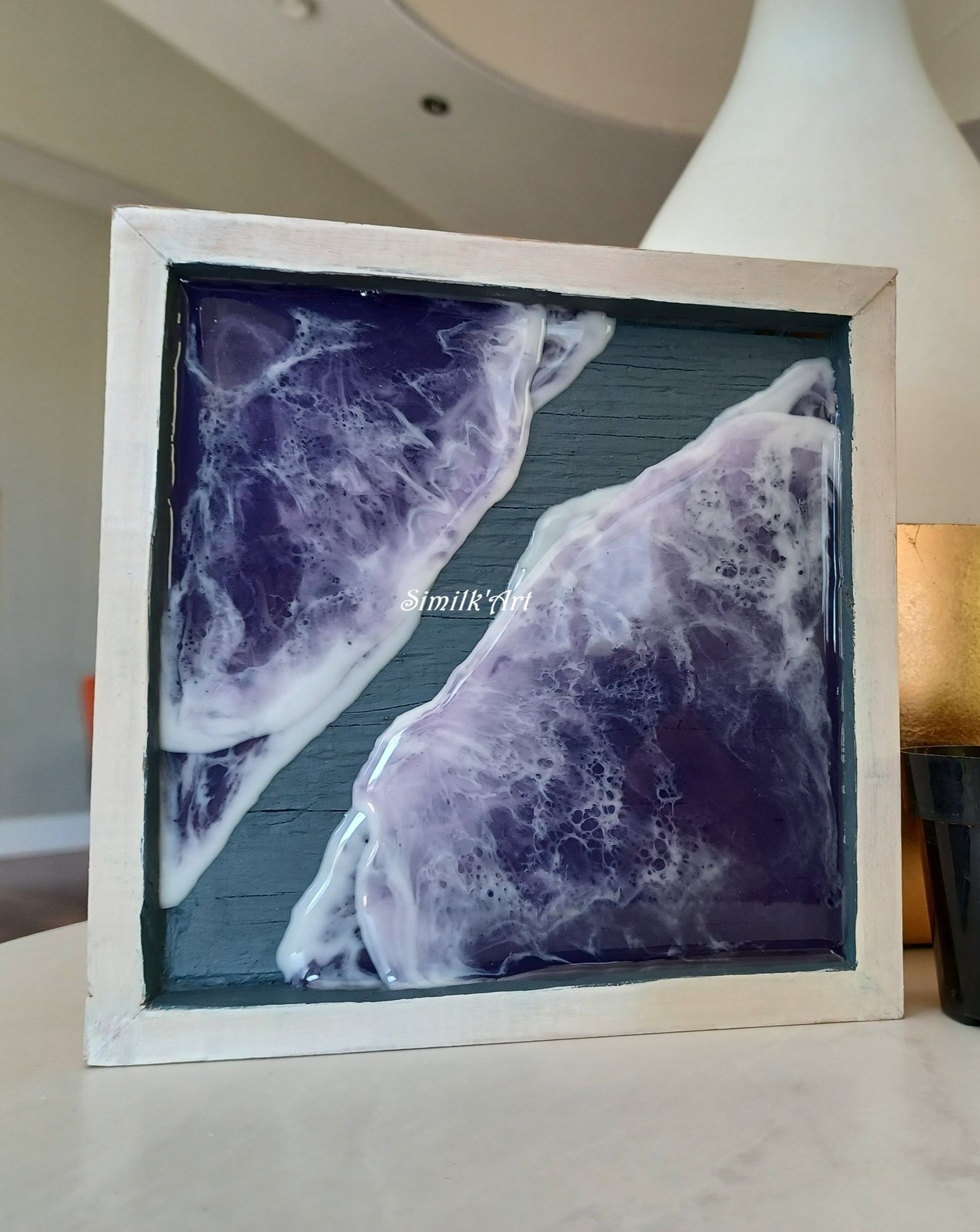 Square shaped art piece made of resin featuring purple ocean and white waves within a white wooden frame.