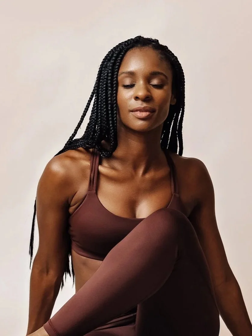 A woman with long braided hair in a brown sports bra and matching leggings, sitting with her eyes closed against a plain white background.