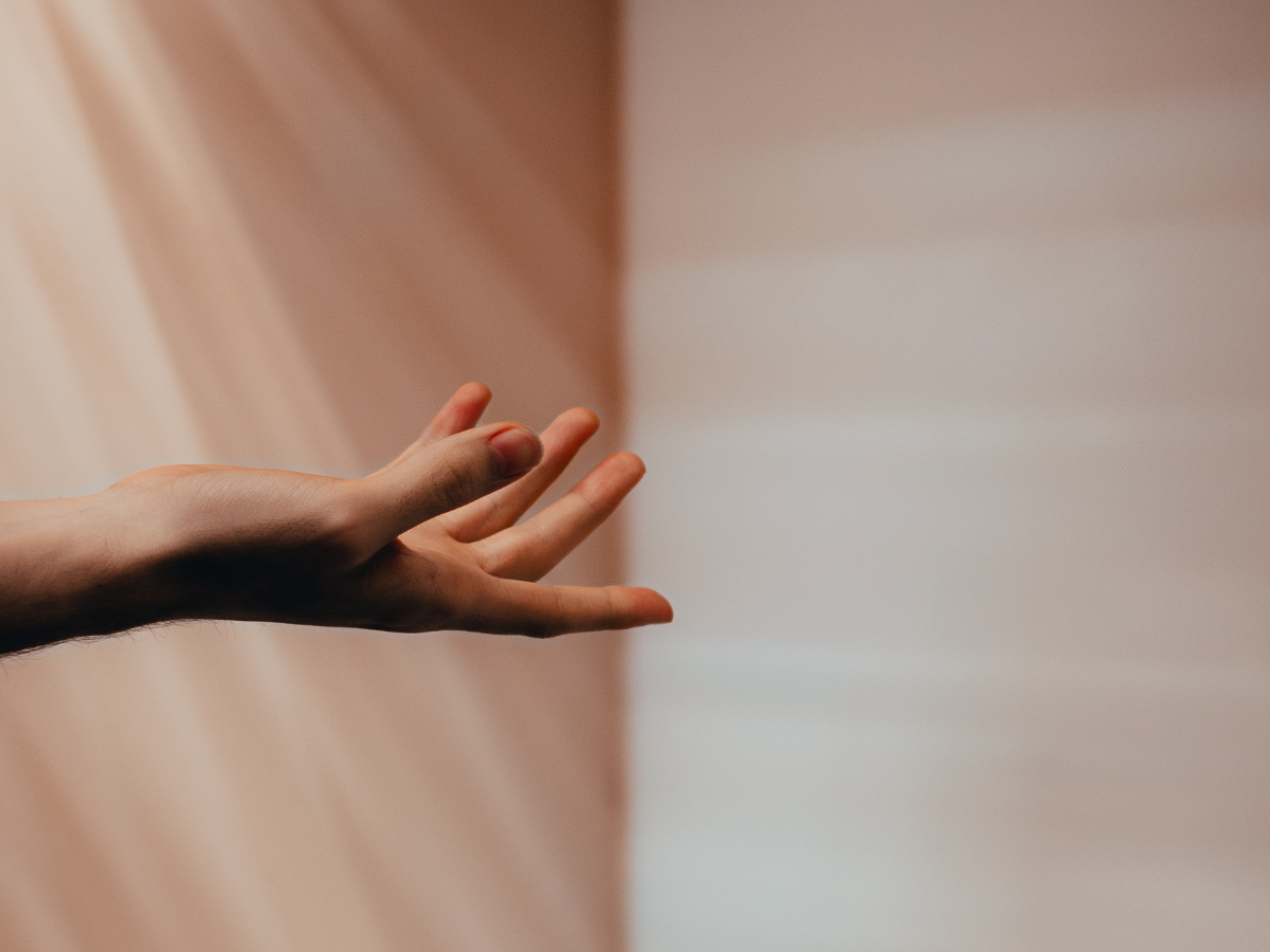 A person's hand with fingers extended, reaching out towards a wall with light and shadow.