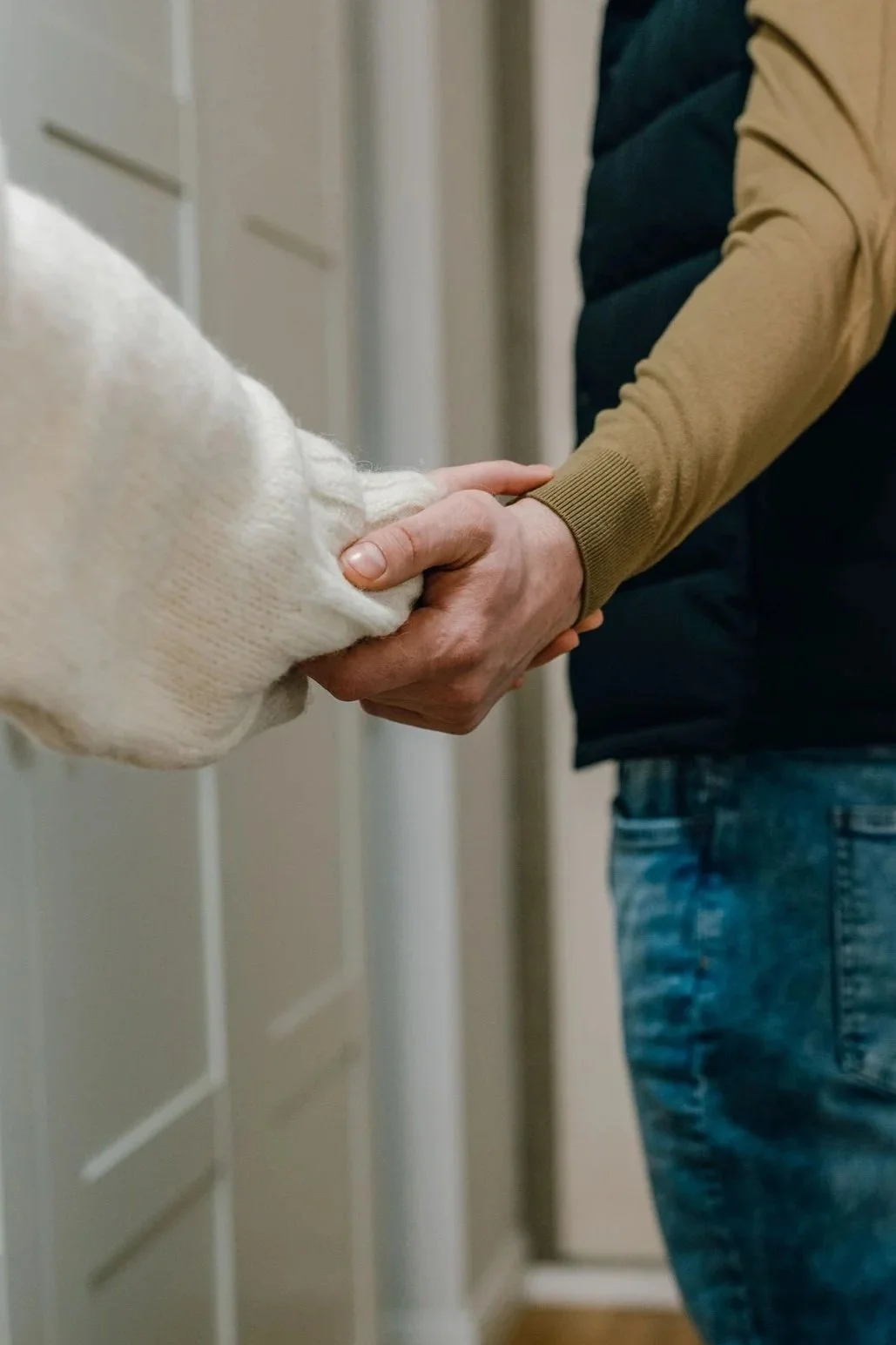 Two people holding hands, one in a white sweater and the other in a tan and black jacket, in an indoor setting.