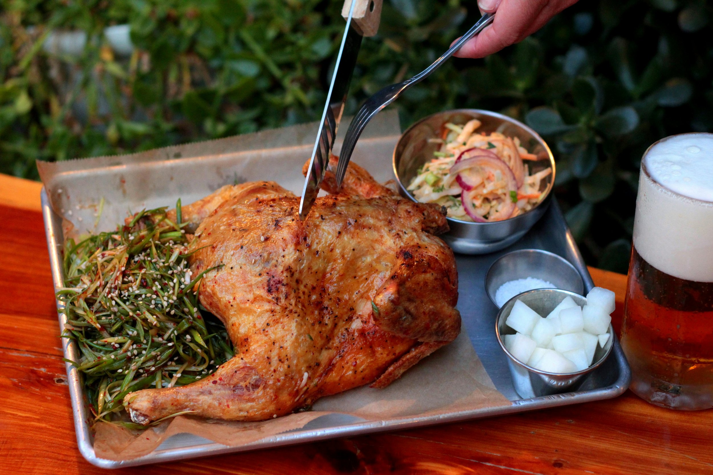 Roasted chicken served with salad, coleslaw, diced onions, salt, and beer on a wooden table.