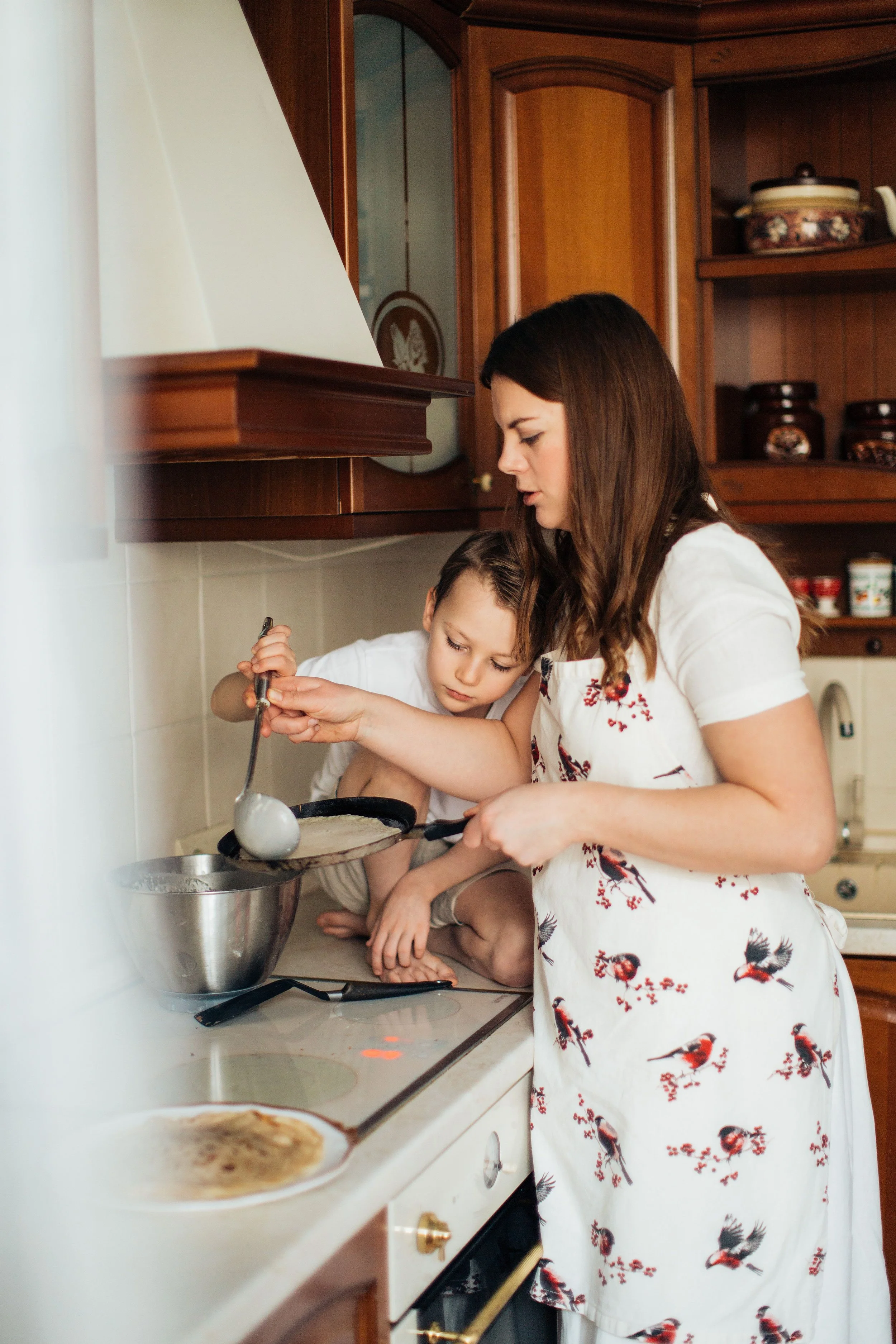 A woman and a young girl cooking together in a kitchen. The woman is wearing a white dress with a bird pattern. The girl is leaning over a bowl, and they appear to be focused on cooking or preparing food.