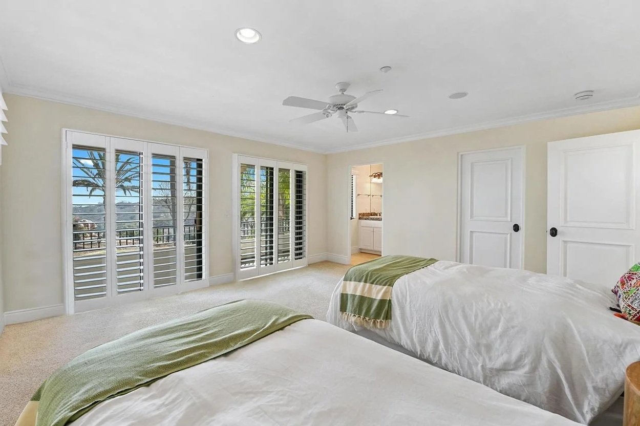 Bedroom with two beds, white linens, green blankets, and colorful pillows, large window with sliding glass doors with plantation shutters, balcony view with trees, ceiling fan, door to a bathroom.