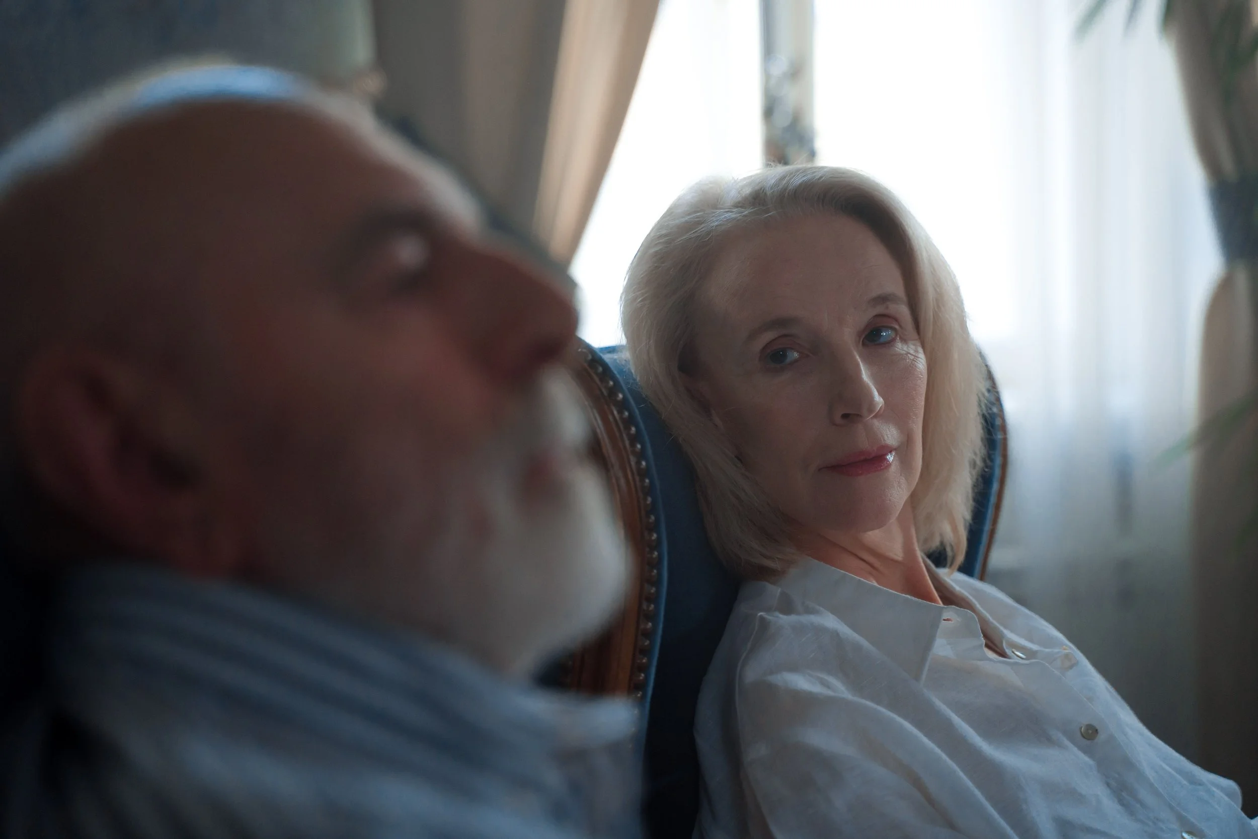 A woman with blonde hair and blue eyes sitting on a train, looking towards a sleeping man with a beard and a beanie, with sunlight coming through the window behind them.