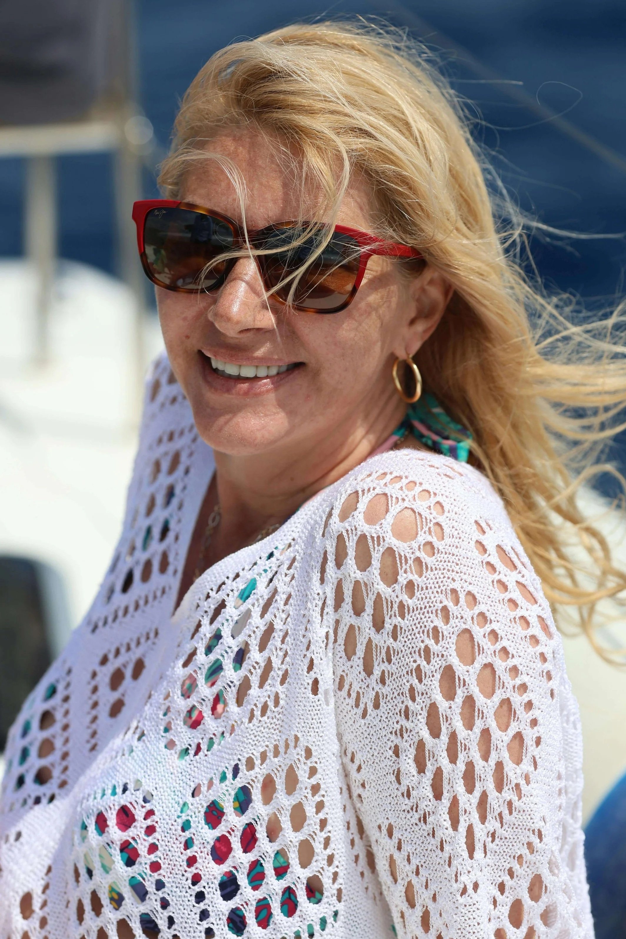 Smiling woman with blonde hair wearing sunglasses and a white crochet top near the water.