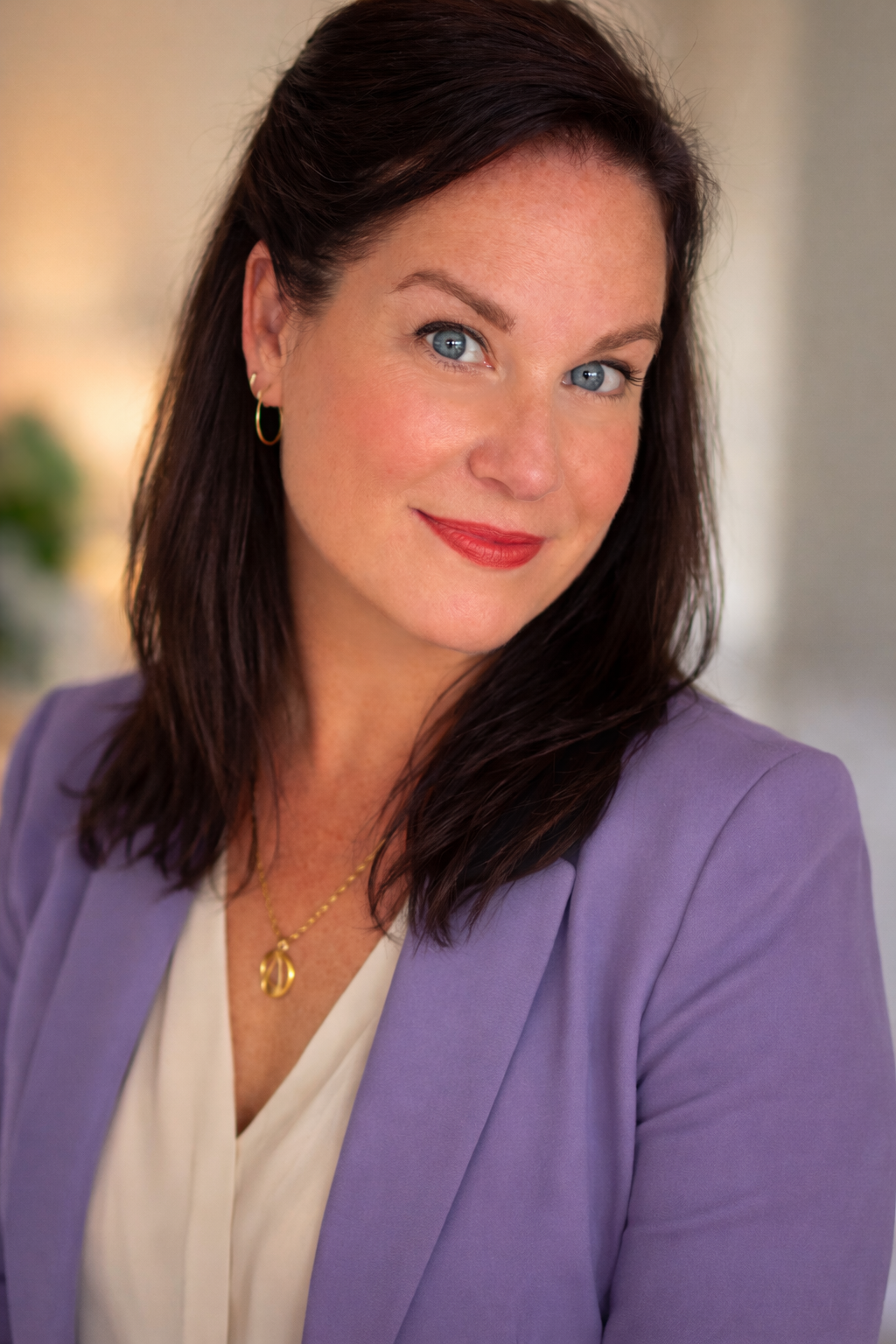 A woman with dark brown hair, blue eyes, and fair skin is smiling at the camera. She is dressed in a light purple blazer over a white top and is wearing gold jewelry, including earrings and a necklace. The background is softly blurred.