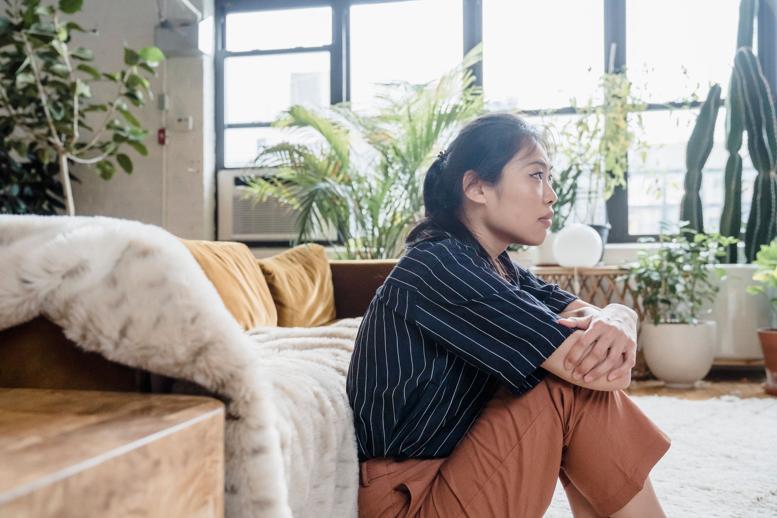 A woman sitting on the floor inside a room with large windows and plenty of indoor plants, wearing a striped shirt and brown pants.