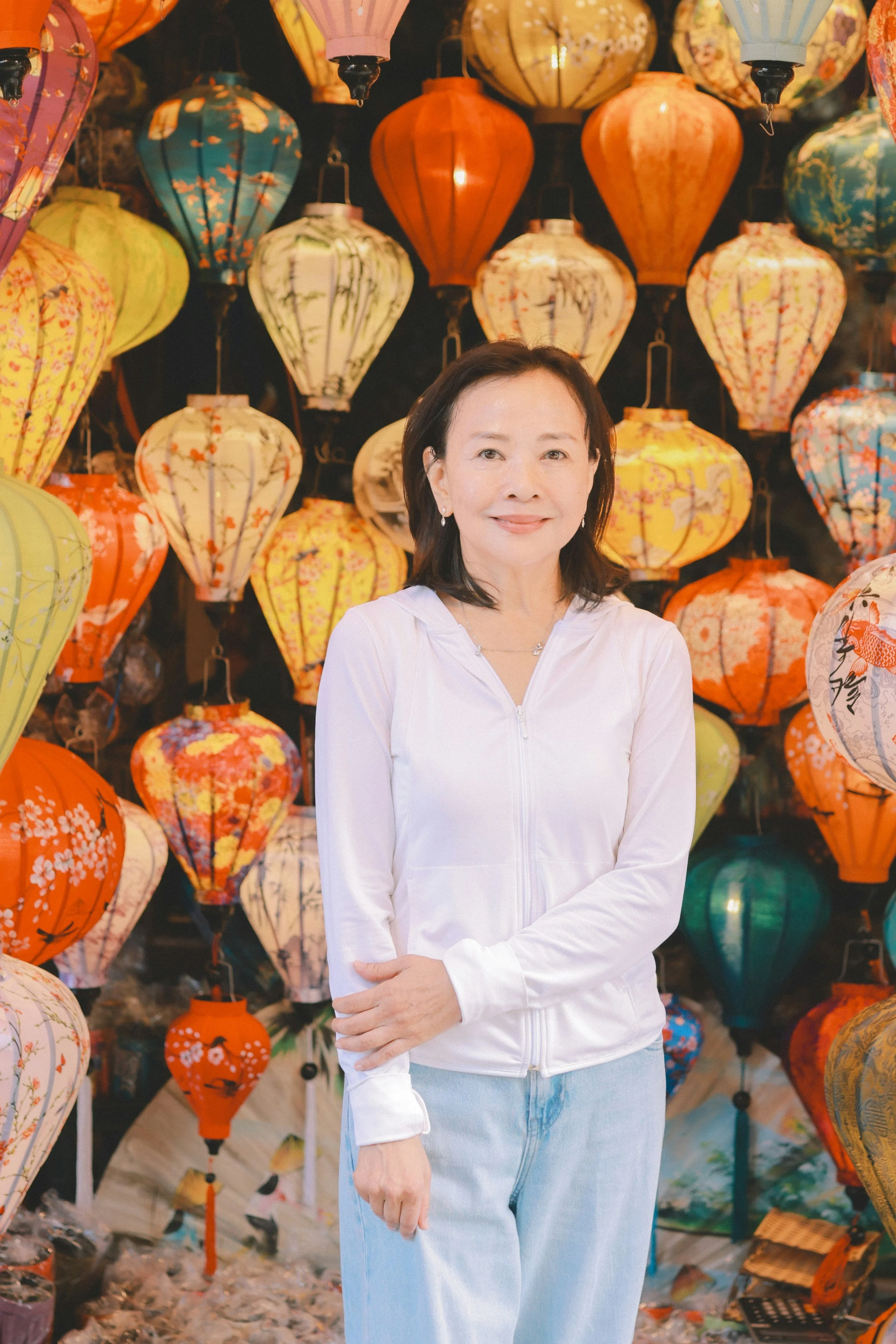 A woman with shoulder-length dark hair is standing in front of a background filled with colorful paper lanterns at night, wearing a white zip-up jacket and light blue jeans, smiling at the camera.