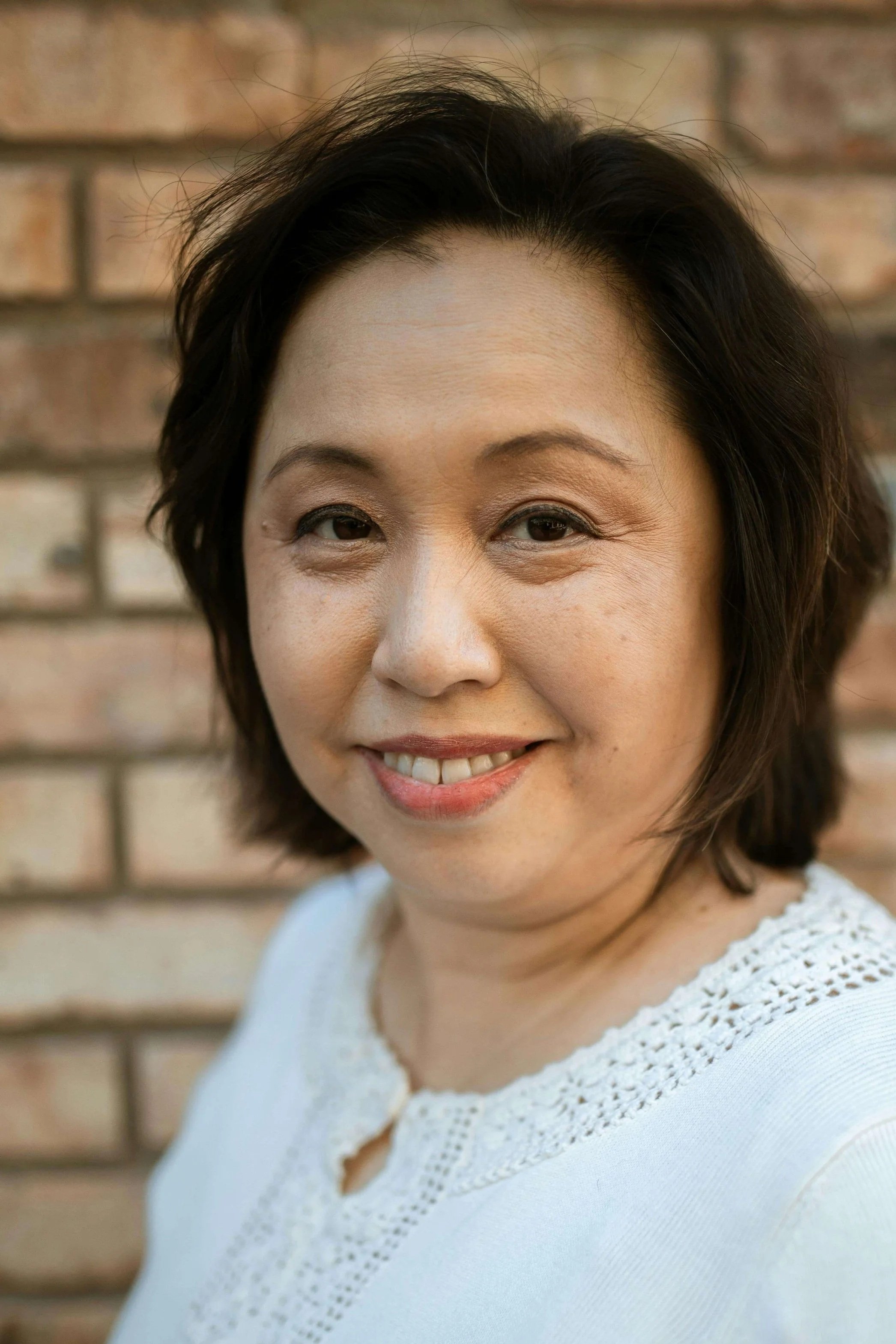 A woman with shoulder-length dark hair smiling in front of a brick wall, wearing a white top with lace details.