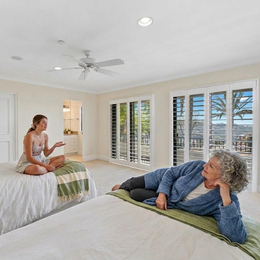 A young woman sitting cross-legged on a bed, speaking to an older woman lying on another bed, in a bright room with large windows and plantation shutters.