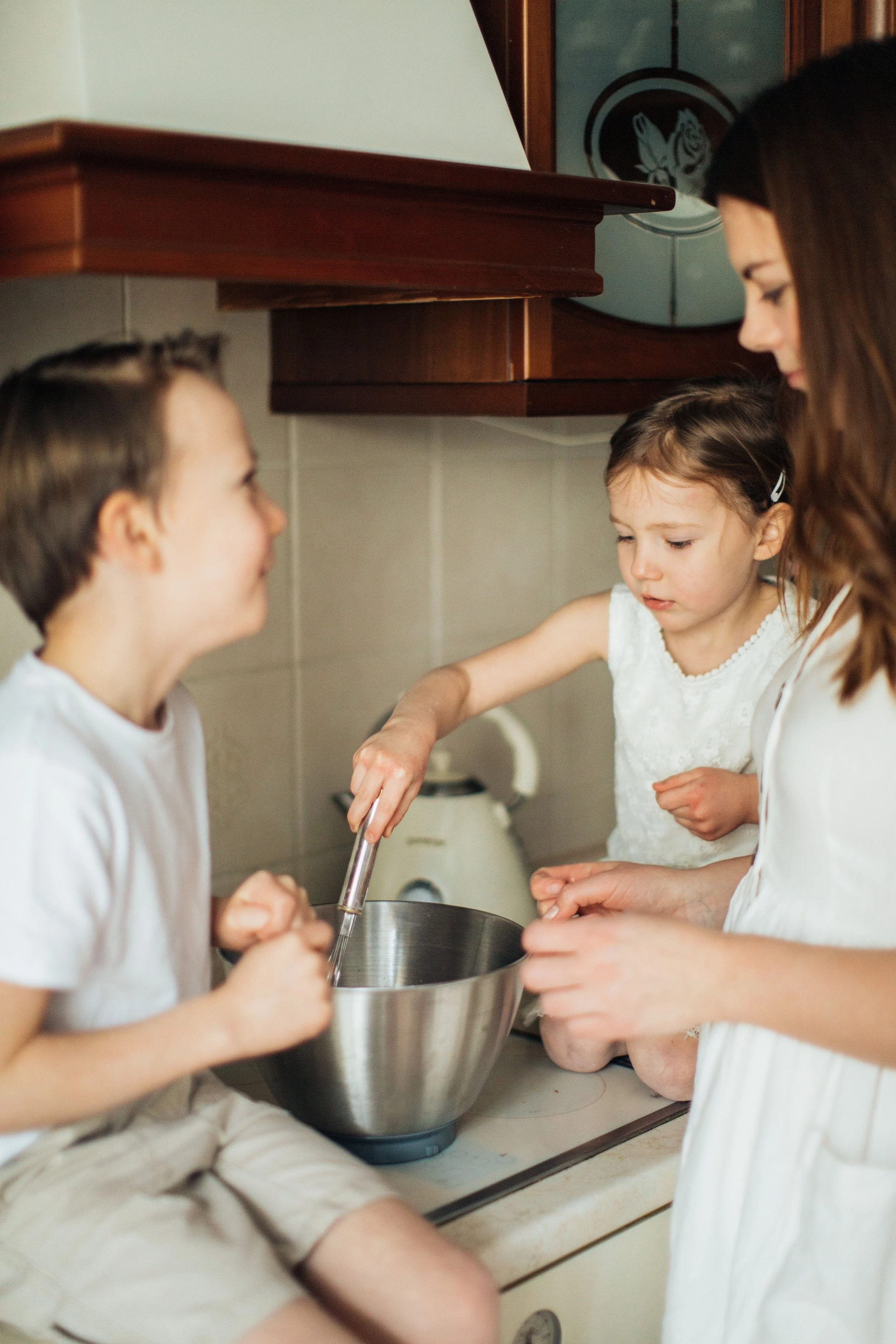 A woman and two children, a boy and a girl, are in a kitchen. The girl is pouring ingredients into a large metal mixing bowl, and the boy is watching, holding a whisk. The woman is standing close, supervising them.